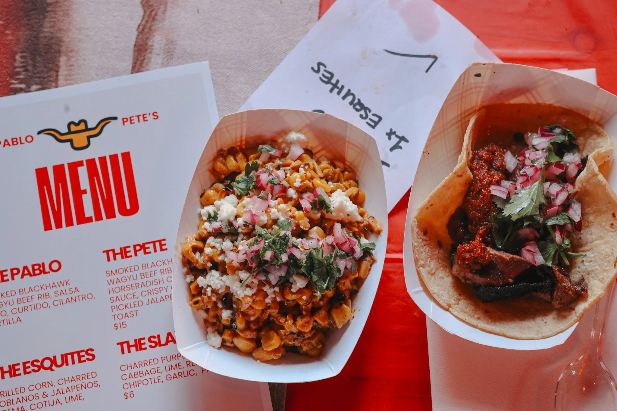 Two dishes of Mexican food, including a taco with meat and toppings and a serving of spicy beans with cheese and herbs on a paper tray, along with a menu and a handwritten note on paper.