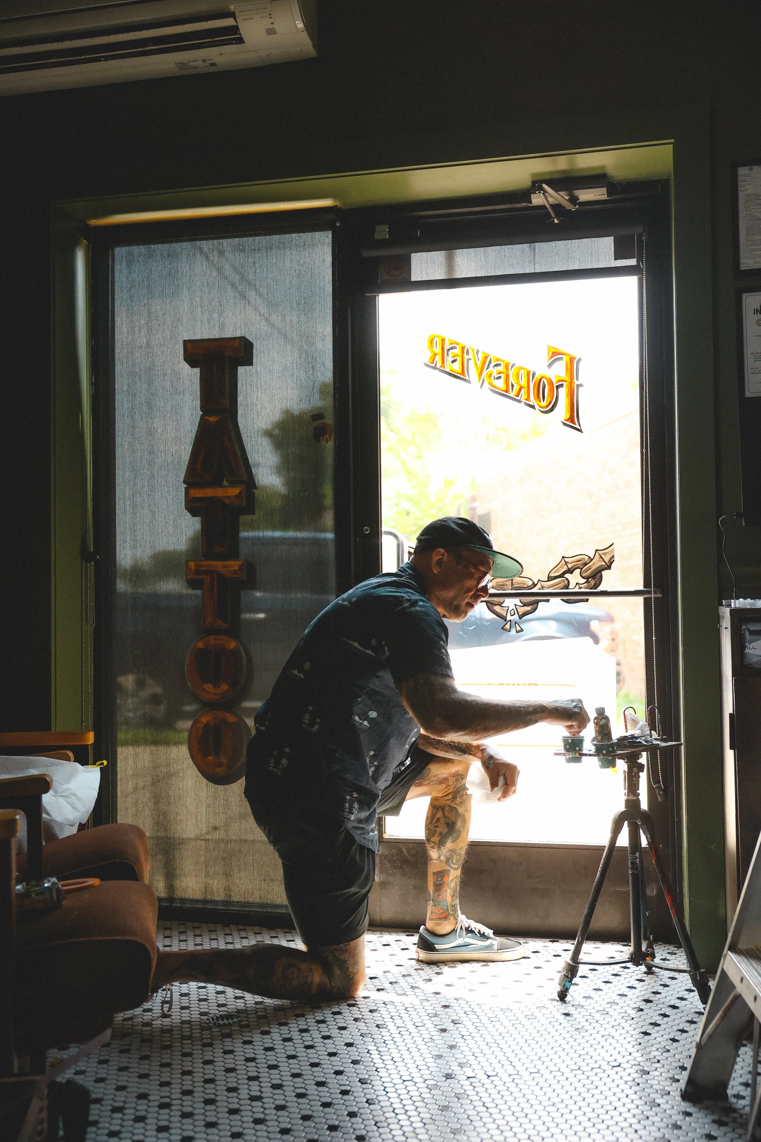 A man with tattoos kneeling on one knee inside a tattoo studio, working on a tattoo on another person's leg. The studio has a glass door with the word "FOREVER" written on it, and artwork is visible on the door and around the room.