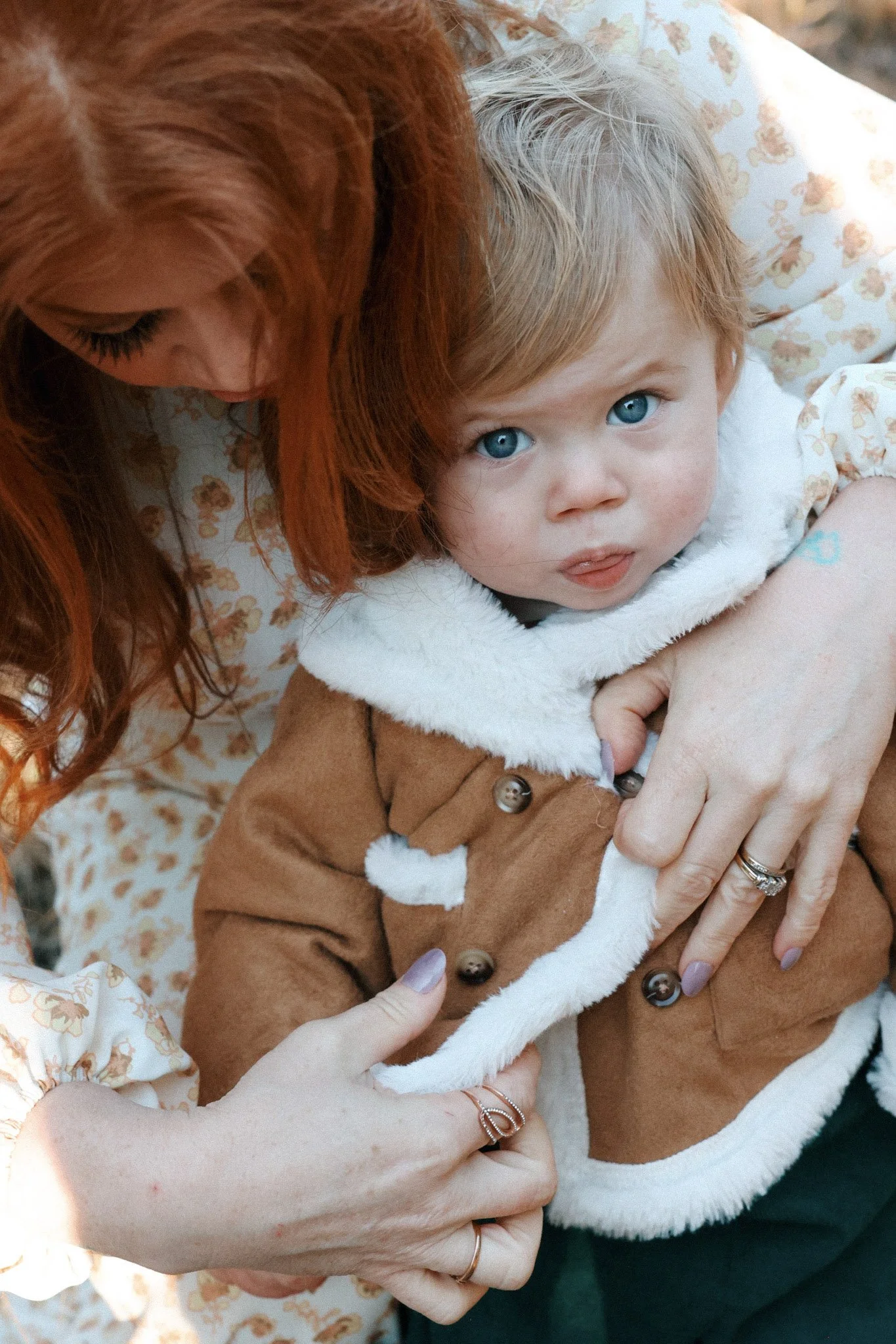 A close-up of a young child with blue eyes being held by an adult woman with red hair. The child is dressed in a brown coat with white fur trim, and the woman is wearing a floral-patterned top.