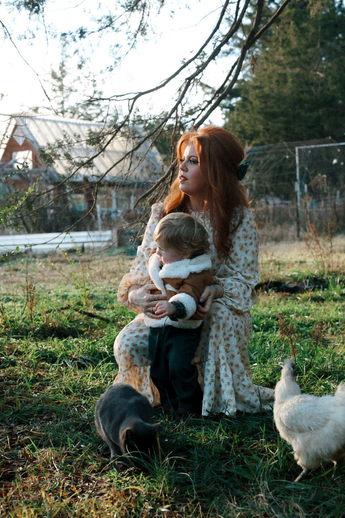 A woman with red hair holding a small child in an outdoor setting with chickens and a cat on the grass.