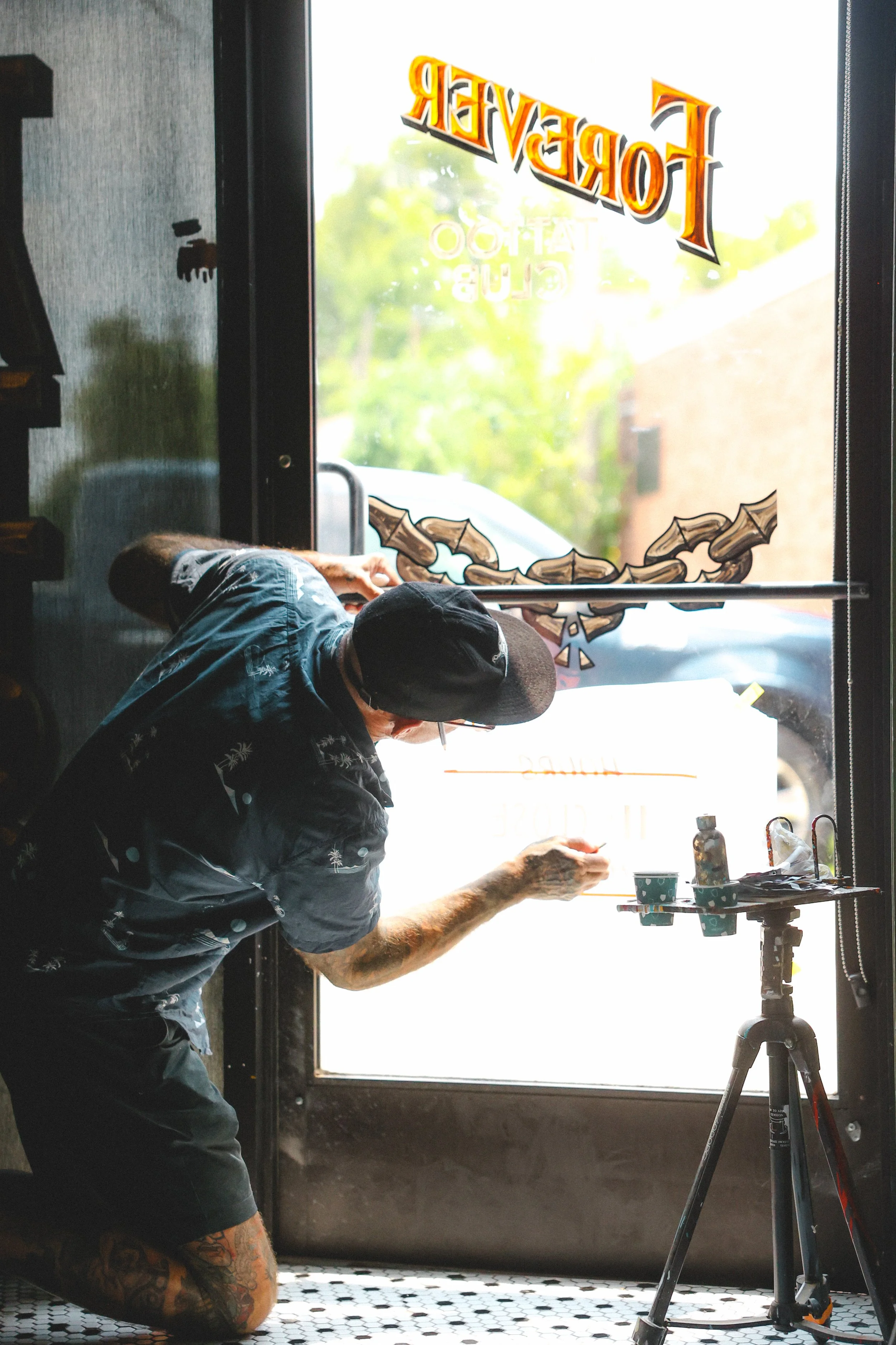 A man with tattoos, wearing a hat and glasses, is designing or painting on a glass door of a store, with outdoor scenery visible through the door.