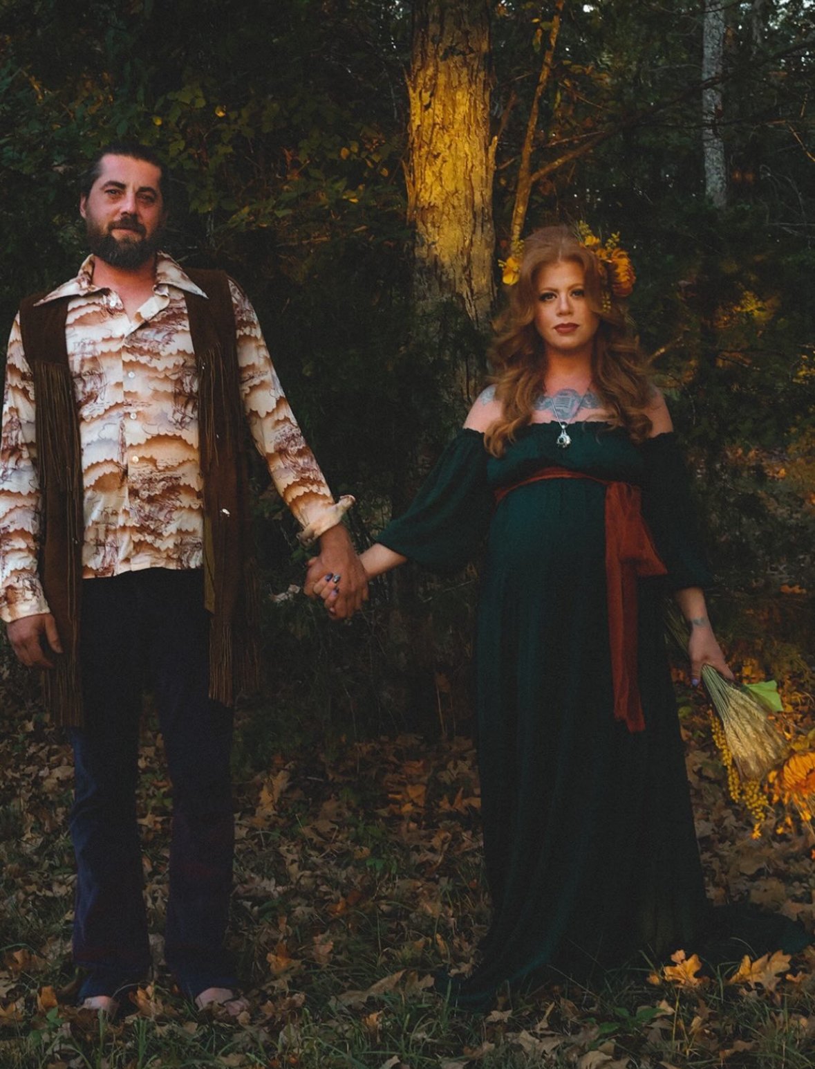 A couple holding hands outdoors in a wooded area during sunset, with autumn leaves on the ground. The man has a beard and wears a patterned shirt with fringe vest, while the woman has long hair, floral earrings, and wears a dark off-shoulder dress wi