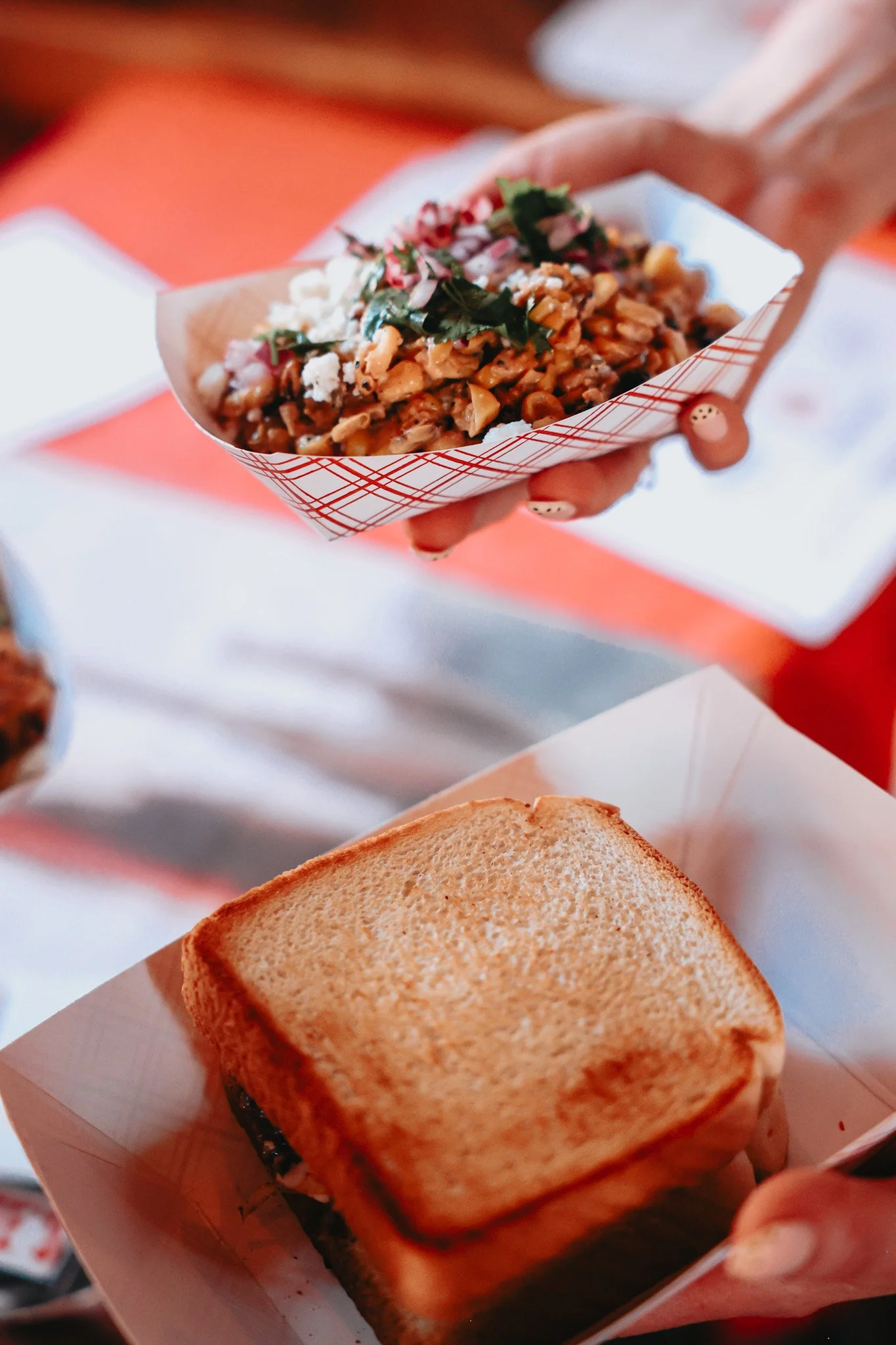 Hand holding a paper tray with a food item topped with herbs and cheese, next to a toasted sandwich on a tray.