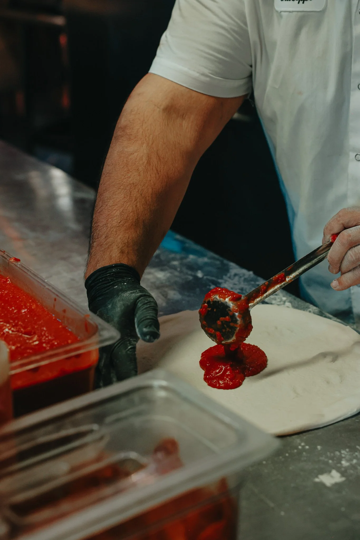 A person spreading tomato sauce onto pizza dough using a ladle at a kitchen counter.