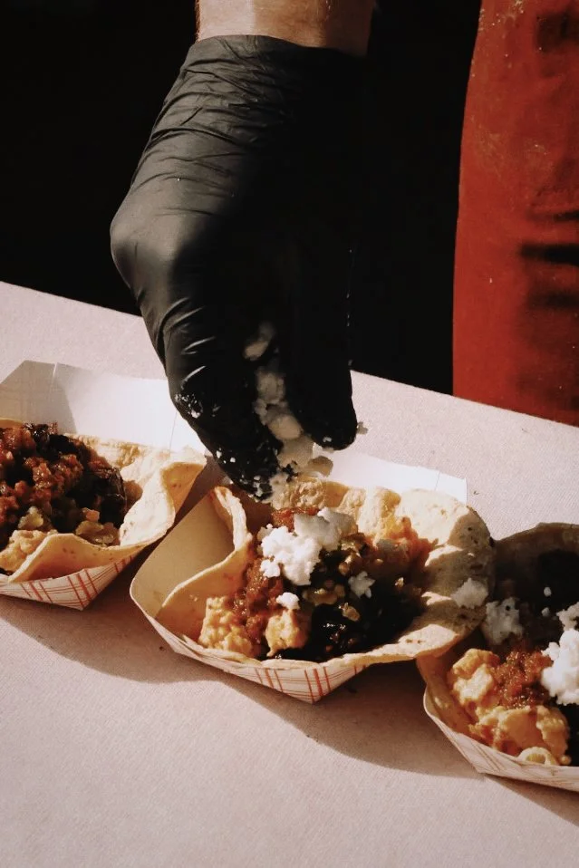 Person wearing a black glove preparing three servings of tacos with ground meat, cheese, and sauce on a white table.