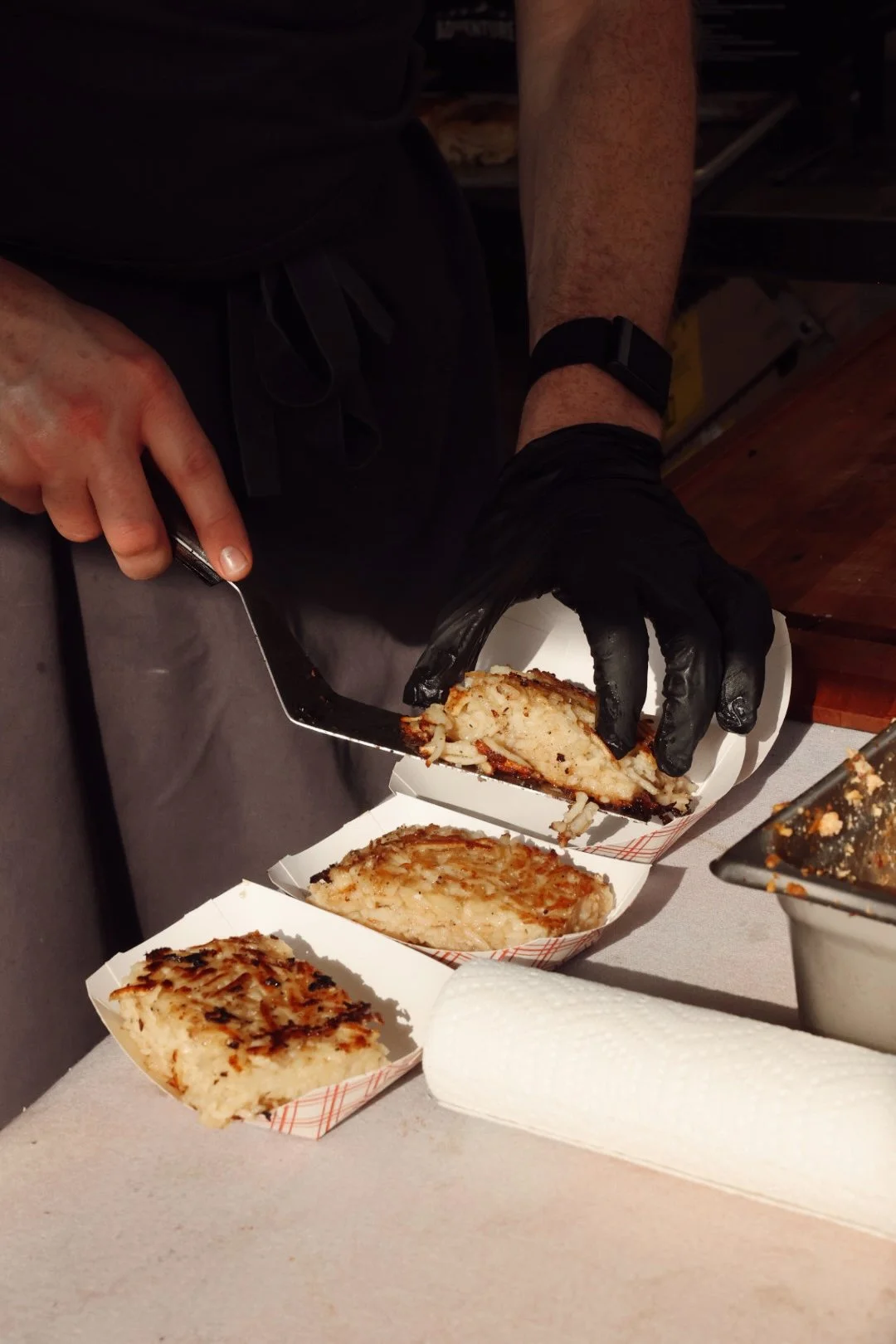 Person wearing black gloves slicing a large piece of grilled or baked chicken or fish into smaller pieces, with trays of similar food on the table.