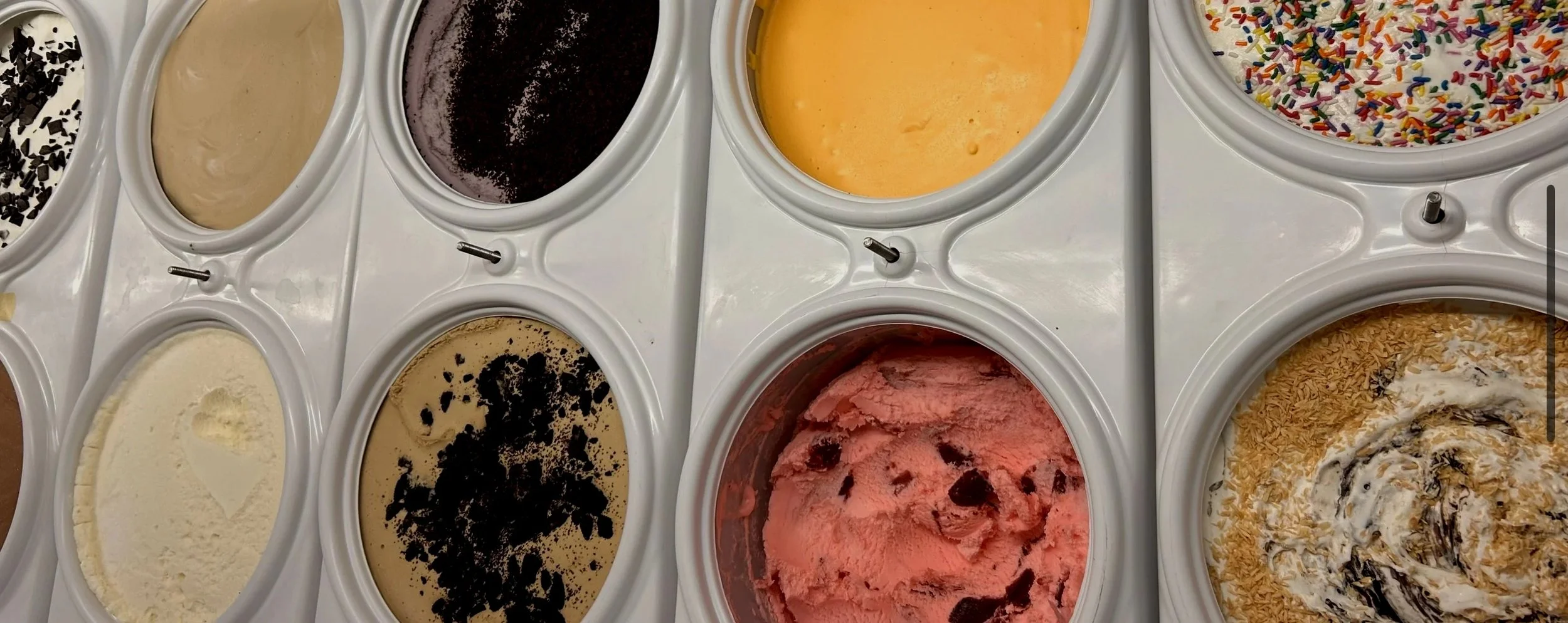 Ice cream flavors in a freezer, including vanilla with chocolate sprinkles, chocolate, mango, rainbow sprinkles, and strawberry with chocolate syrup