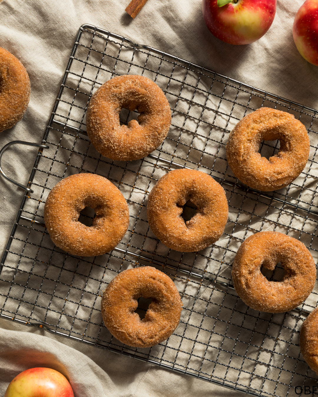 Six apple cider donuts cooling on a wire rack with apples on a light-colored fabric background.