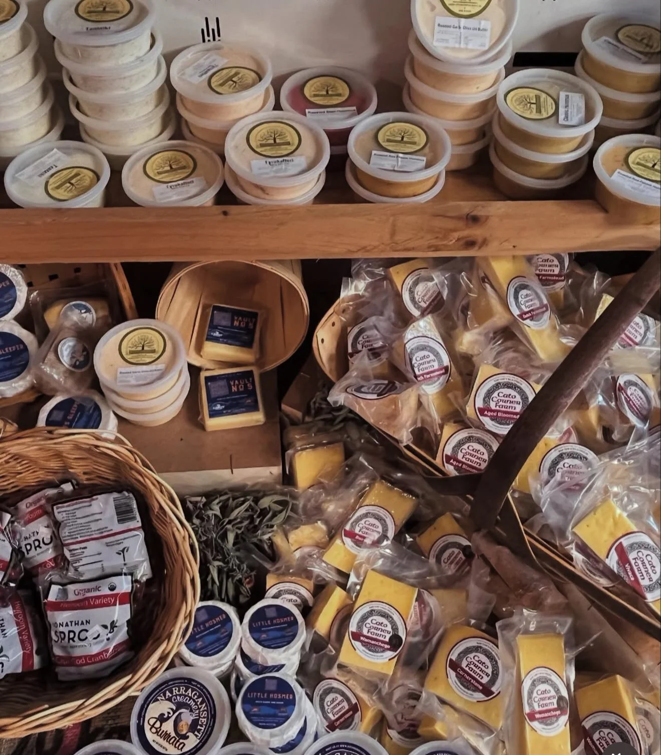 Various types of cheese displayed on shelves and in baskets at a market, including rounds and blocks of cheese packaged in plastic and paper.