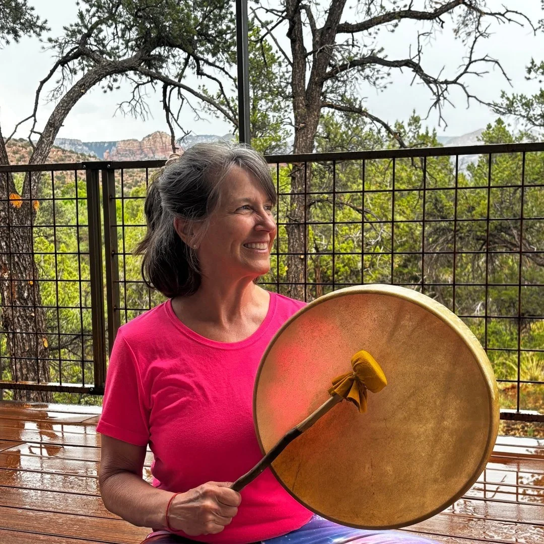 A young man with curly hair standing in a grassy field holding a drum, wearing a plaid jacket and colorful clothing, with an overcast sky in the background.