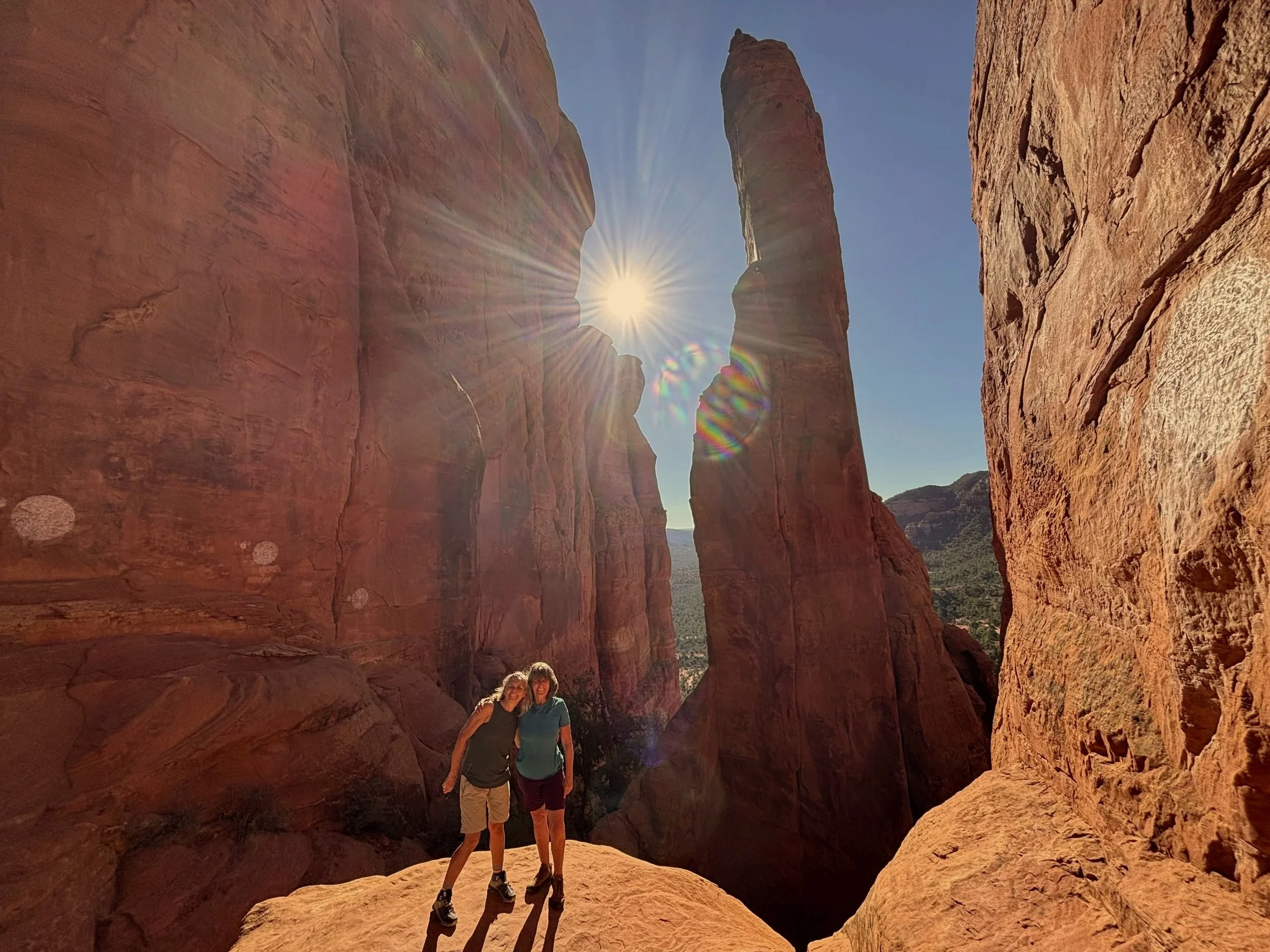 A woman standing between two large red rock formations in a desert landscape, with a distant view of more red rocks and greenery under a partly cloudy sky.