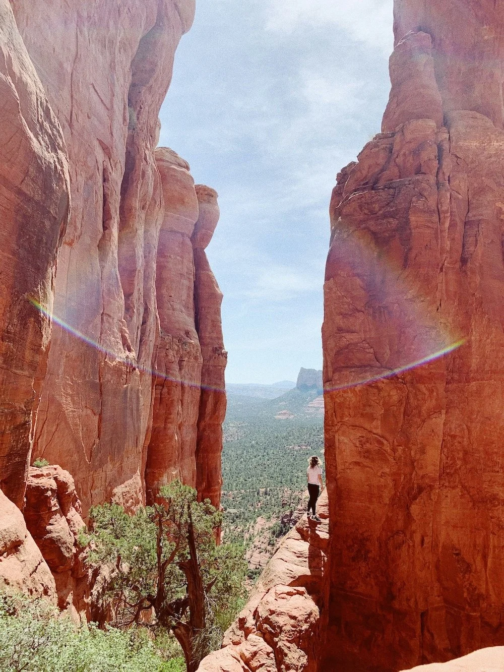 A person standing on a narrow ledge between two tall red rock cliffs in a desert landscape, with a forested valley and distant mesas in the background under a partly cloudy sky.