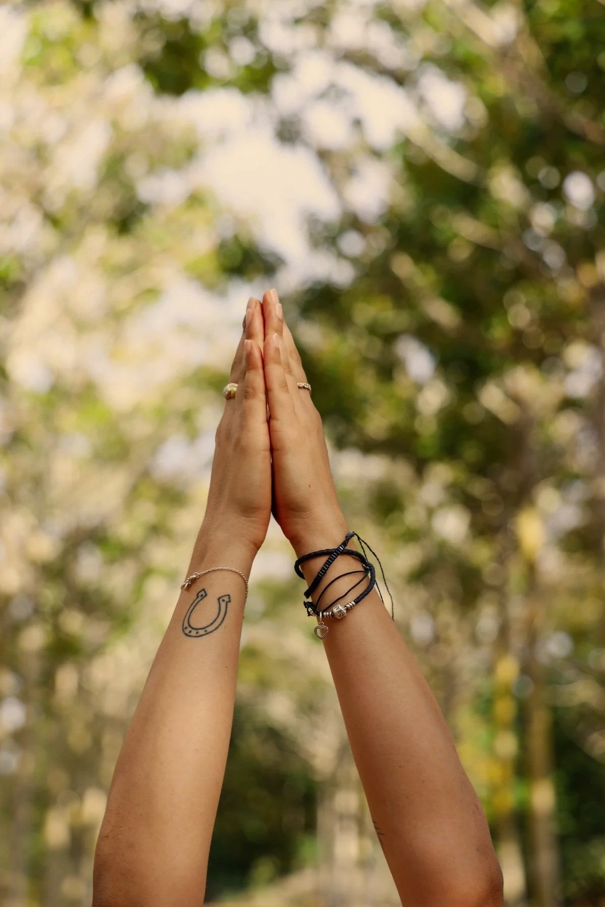Two hands pressed together in a prayer or greeting gesture against a blurred green and blue outdoor background.