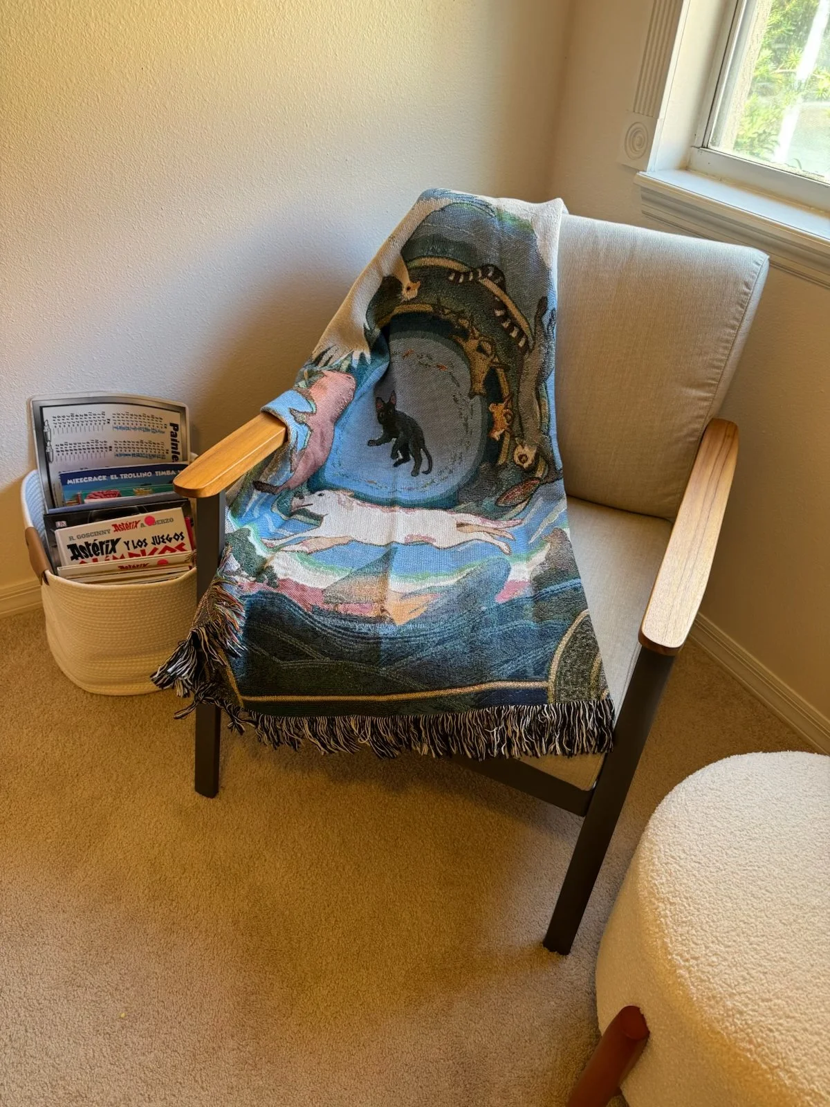 Armchair with a colorful tapestry featuring a black cat, a white dog, and a historical scene; nearby basket with magazines, and a window showing greenery outside.