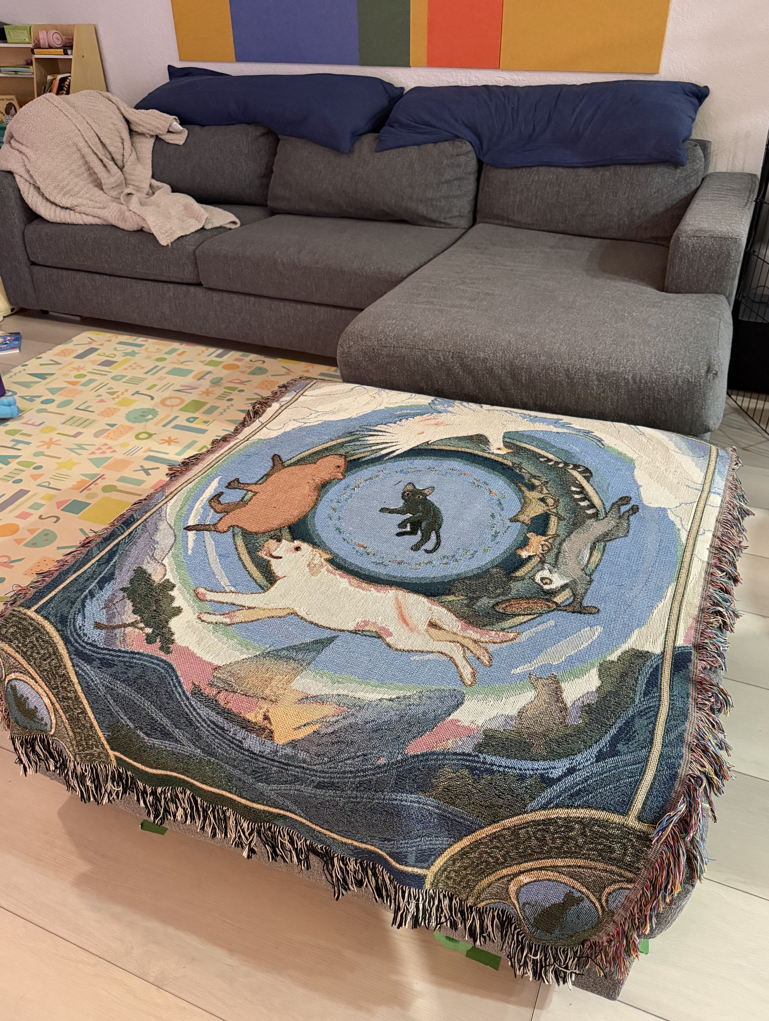 A gray sectional sofa with blue pillows and a blanket on it. In front, there is a colorful tapestry with a cat-themed design featuring cats around a swirling background, his is on a light wood floor, with a colorful abstract art piece on the wall beh