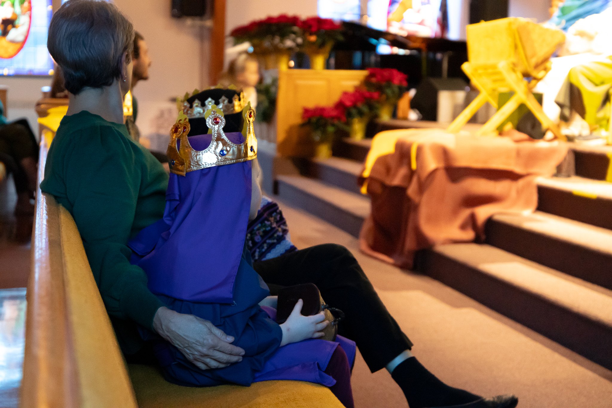 A young girl wearing a princess crown sitting on a bench in a decorated room, possibly a church or community hall, with other people and Christmas poinsettias in the background.