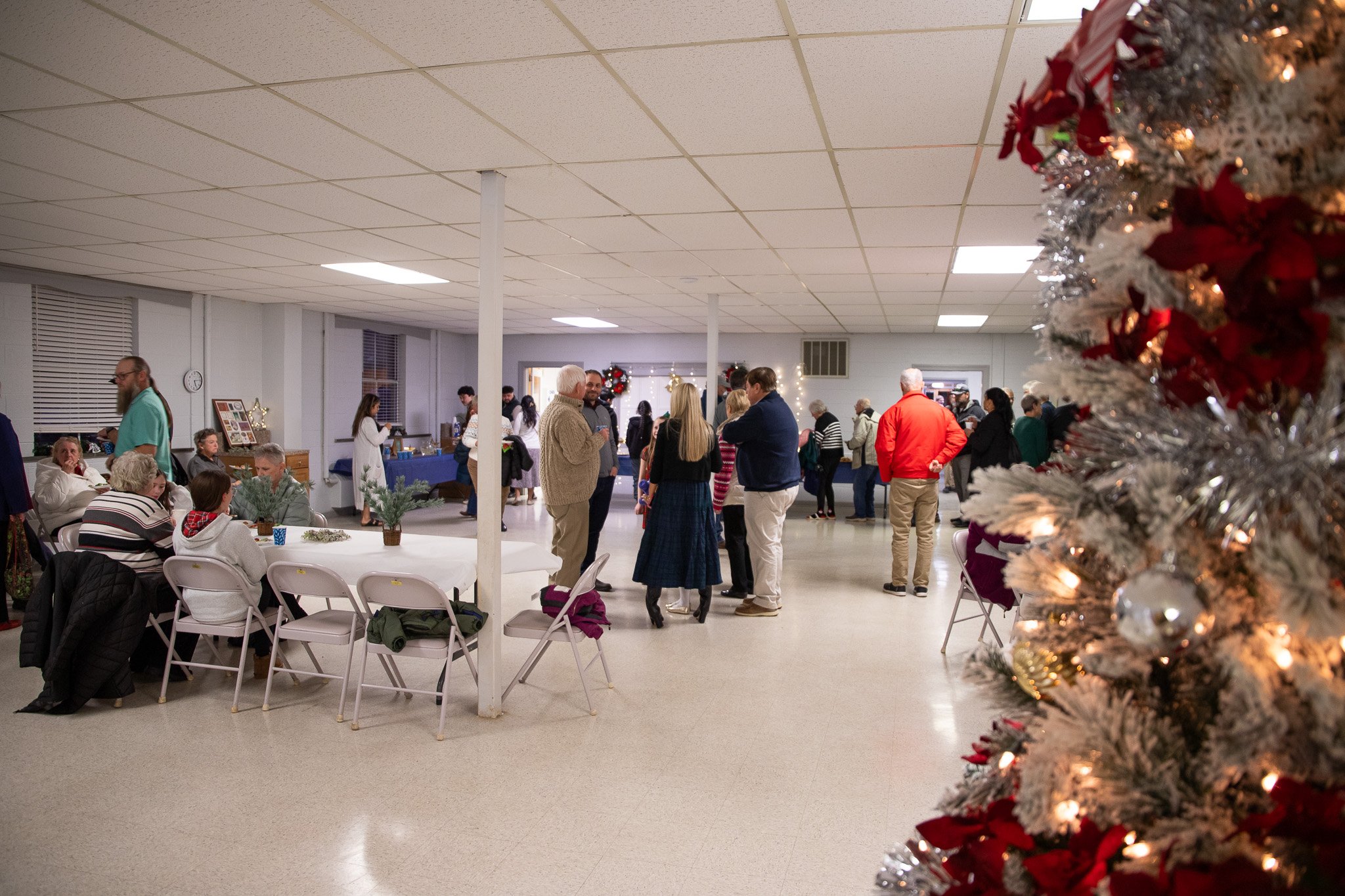 People gathering at a Christmas party in a community hall decorated with wreaths and lights, with a Christmas tree partially visible on the right side of the image.