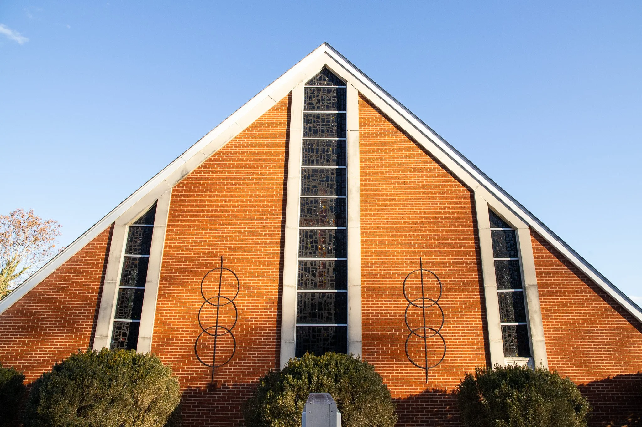 Front view of a brick church with a steep gabled roof, stained glass windows, and metal target-shaped art on the exterior wall, set against a clear blue sky.