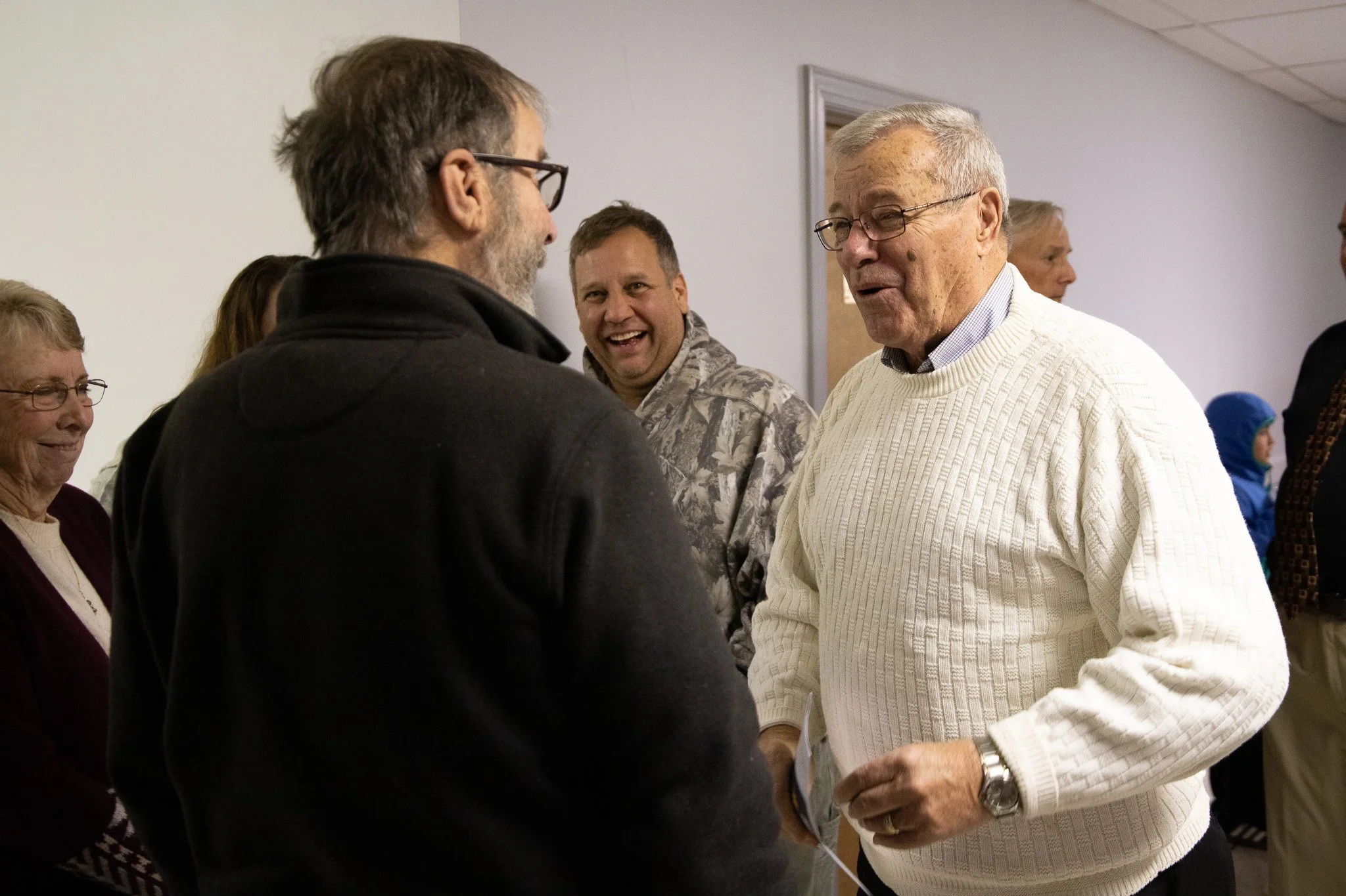 A group of people socializing, including an elderly man in a white sweater and a man in glasses, in an indoor setting.