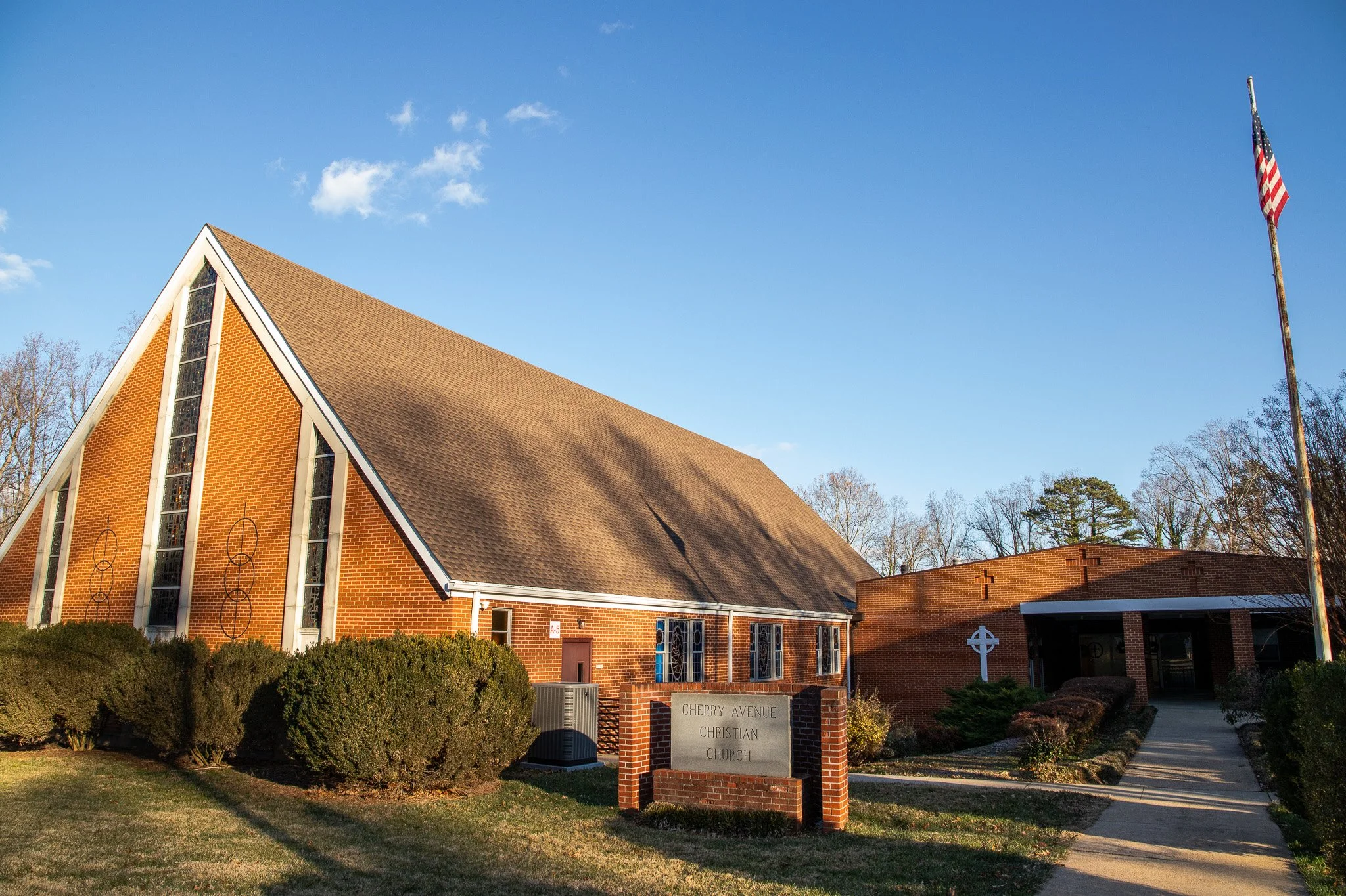 A red brick church with a steep triangular roof and stained glass windows, surrounded by trees and bushes. An American flag is on a tall flagpole to the right. A sign in front identifies it as Cherry Avenue Christian Church.