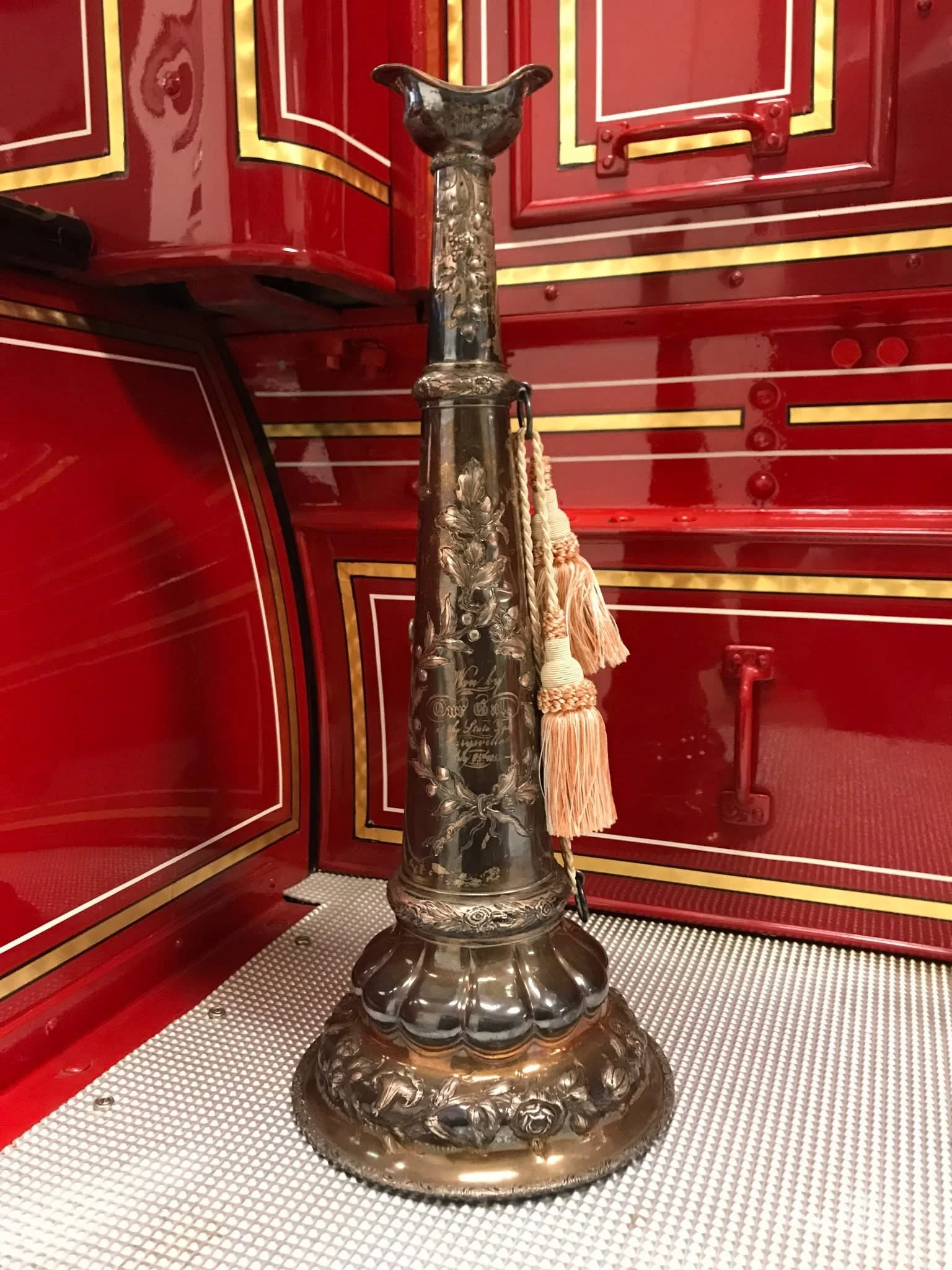 A silver ceremonial mace with floral engravings and a chain with tassels hanging from it, displayed against a glossy red and gold cabinet.