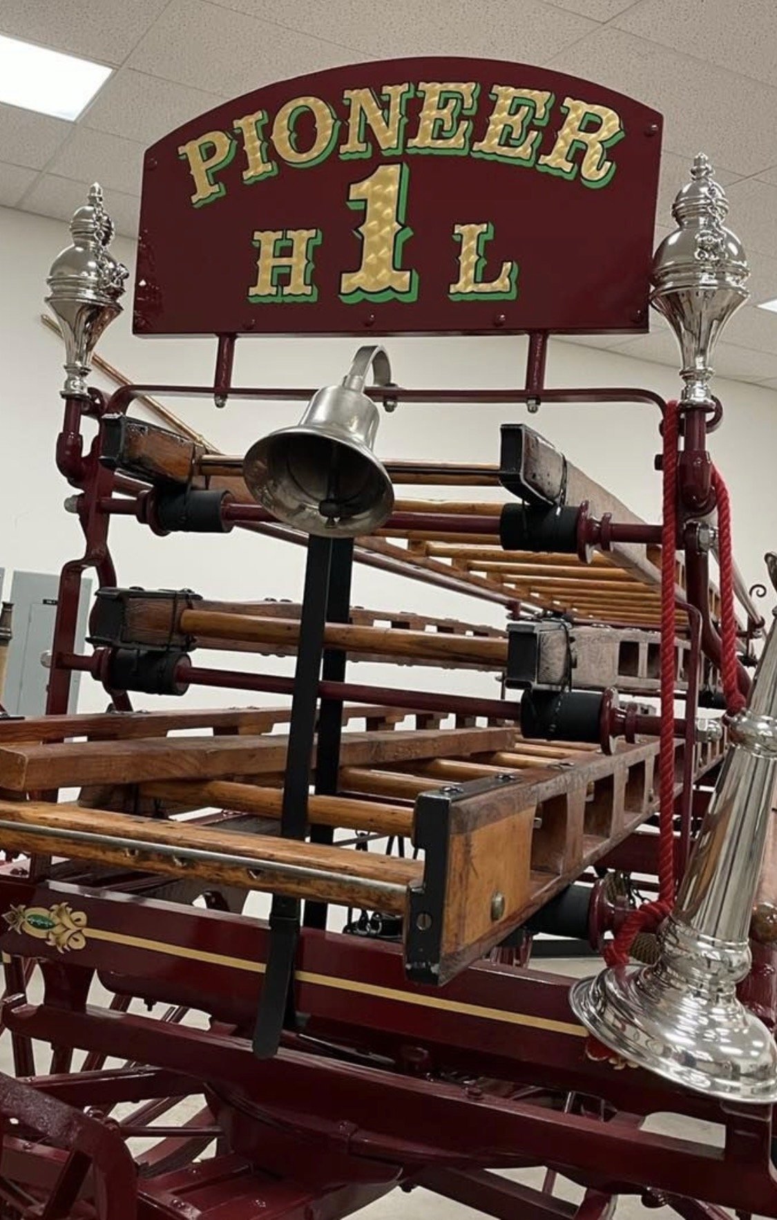 A vintage red fire truck with a wooden ladder and brass fittings, a sign reading 'PIONEER H L 1' at the top, and a large silver horn attached to the side.