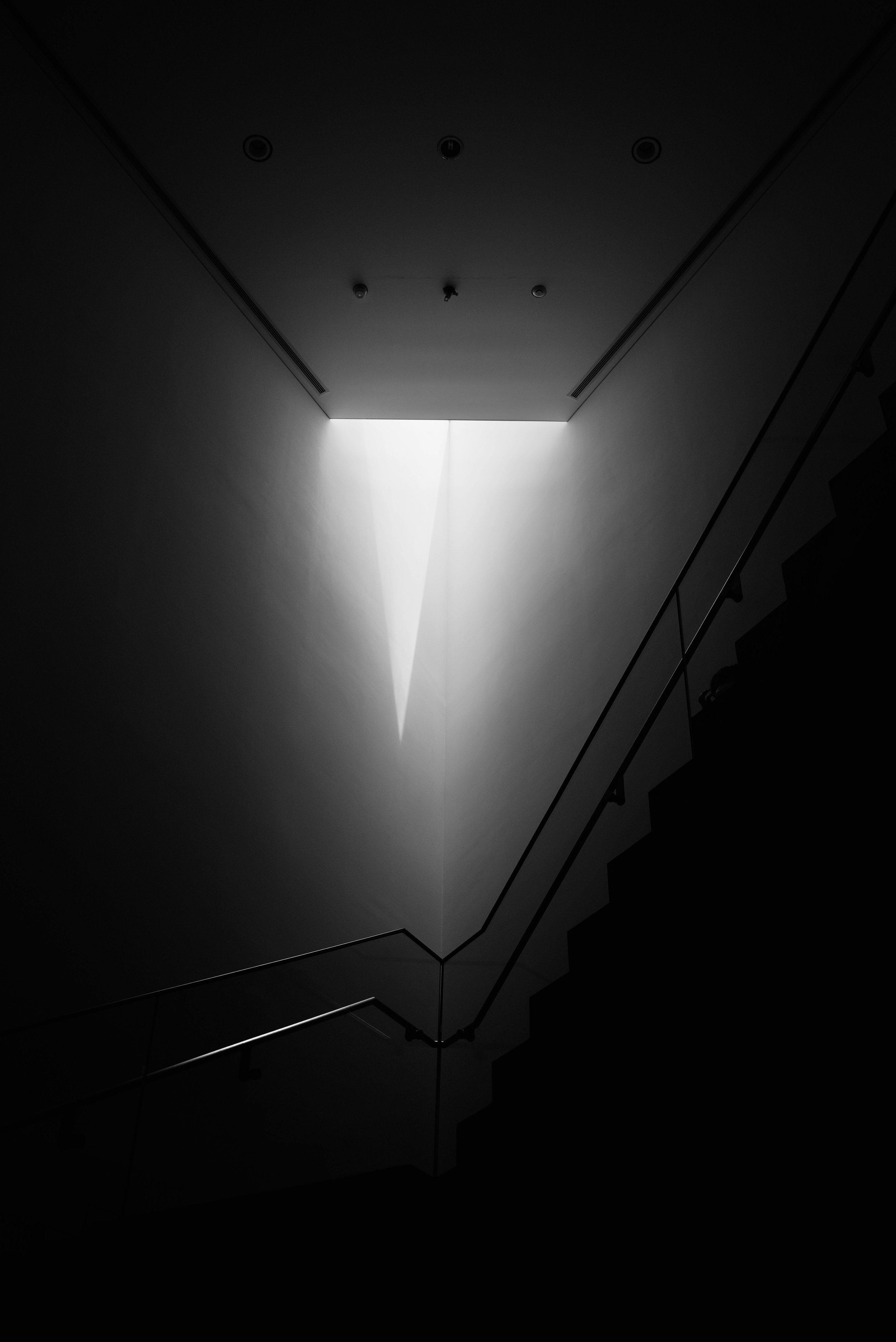 Dark staircase with a minimalistic design, illuminated by a narrow beam of light coming from a skylight overhead.