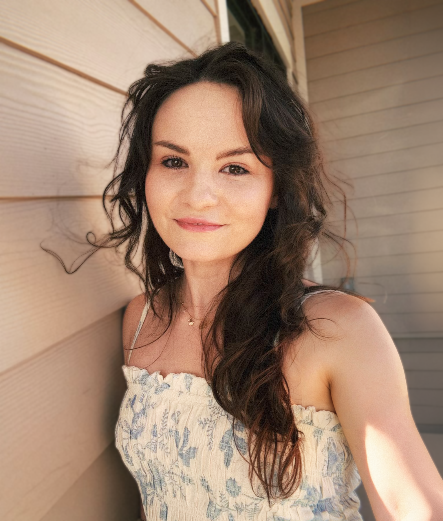 A woman with long, wavy dark hair smiling outdoors beside a wooden wall.