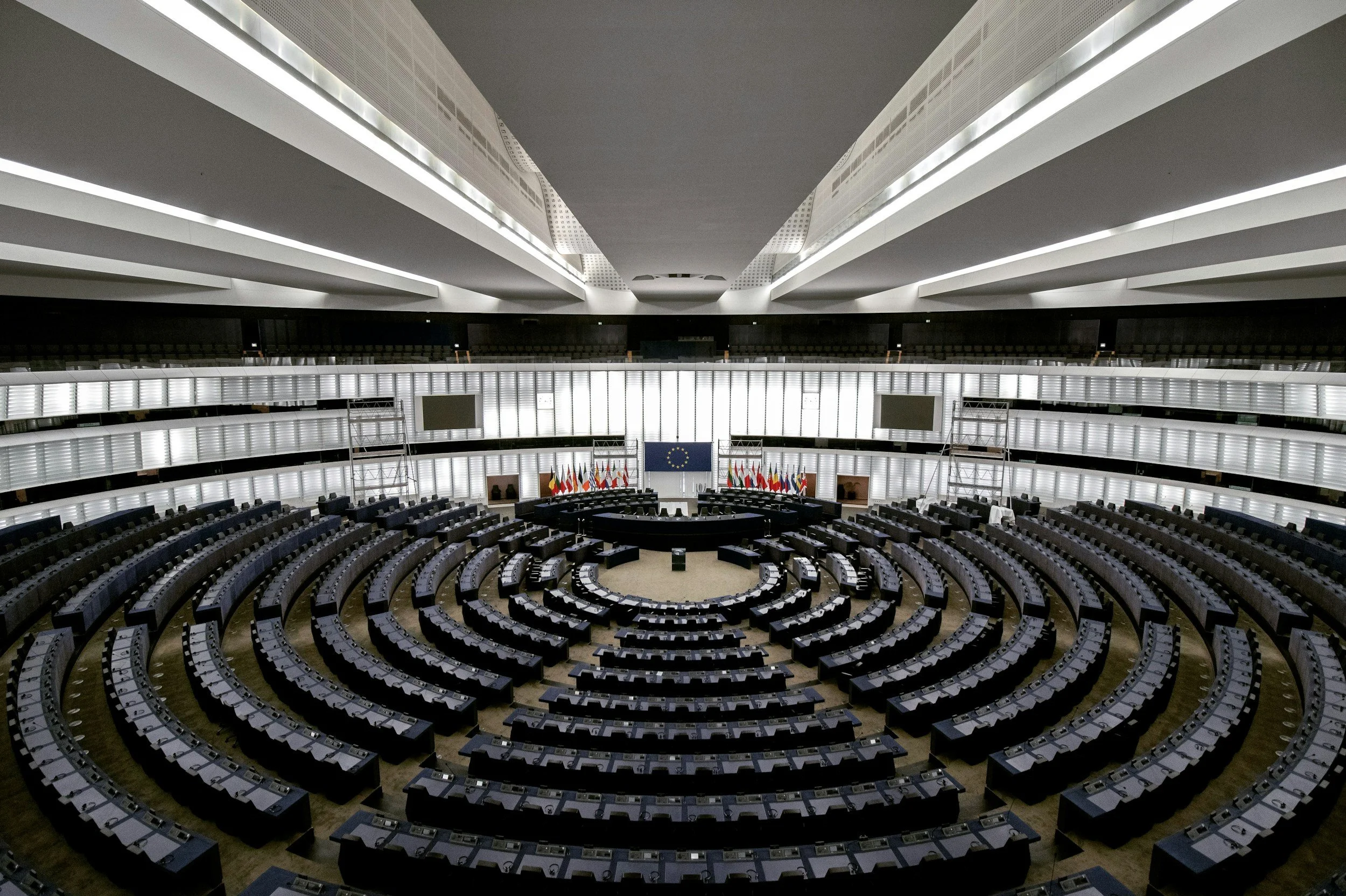 Empty European Parliament chamber with rows of seats arranged in a semi-circle around a central podium, large European Union flag on a screen, and various national flags behind.