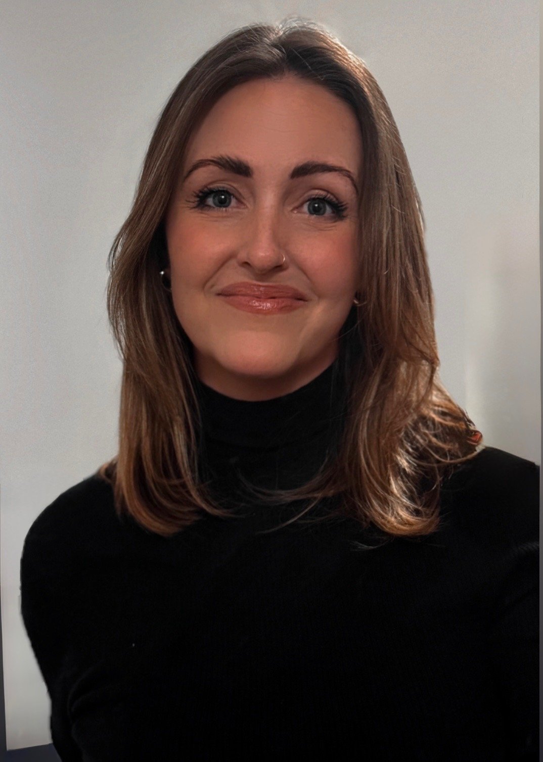 A woman with shoulder-length brown hair, wearing a black turtleneck, smiling, against a plain light-colored background.