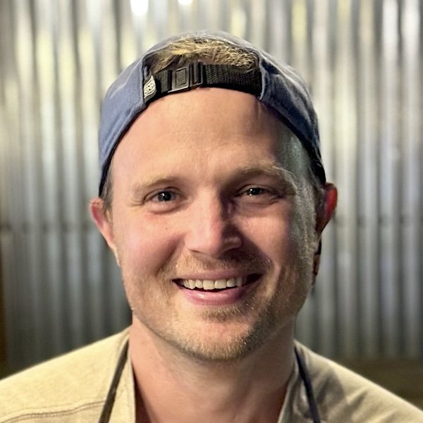 A smiling man wearing a backward gray baseball cap, with a blurred metal background.