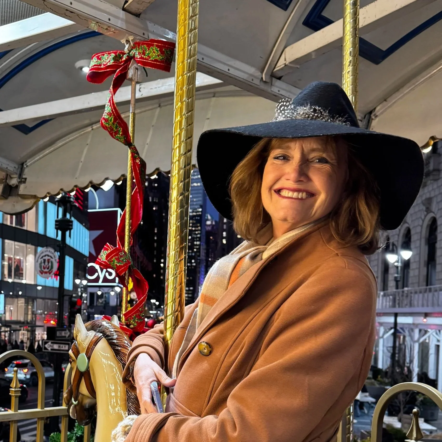 A woman smiling and wearing a large black floppy hat, a beige scarf, and a brown coat while standing near a carousel in an urban setting at night with tall buildings and city lights in the background, decorated for the holiday season.
