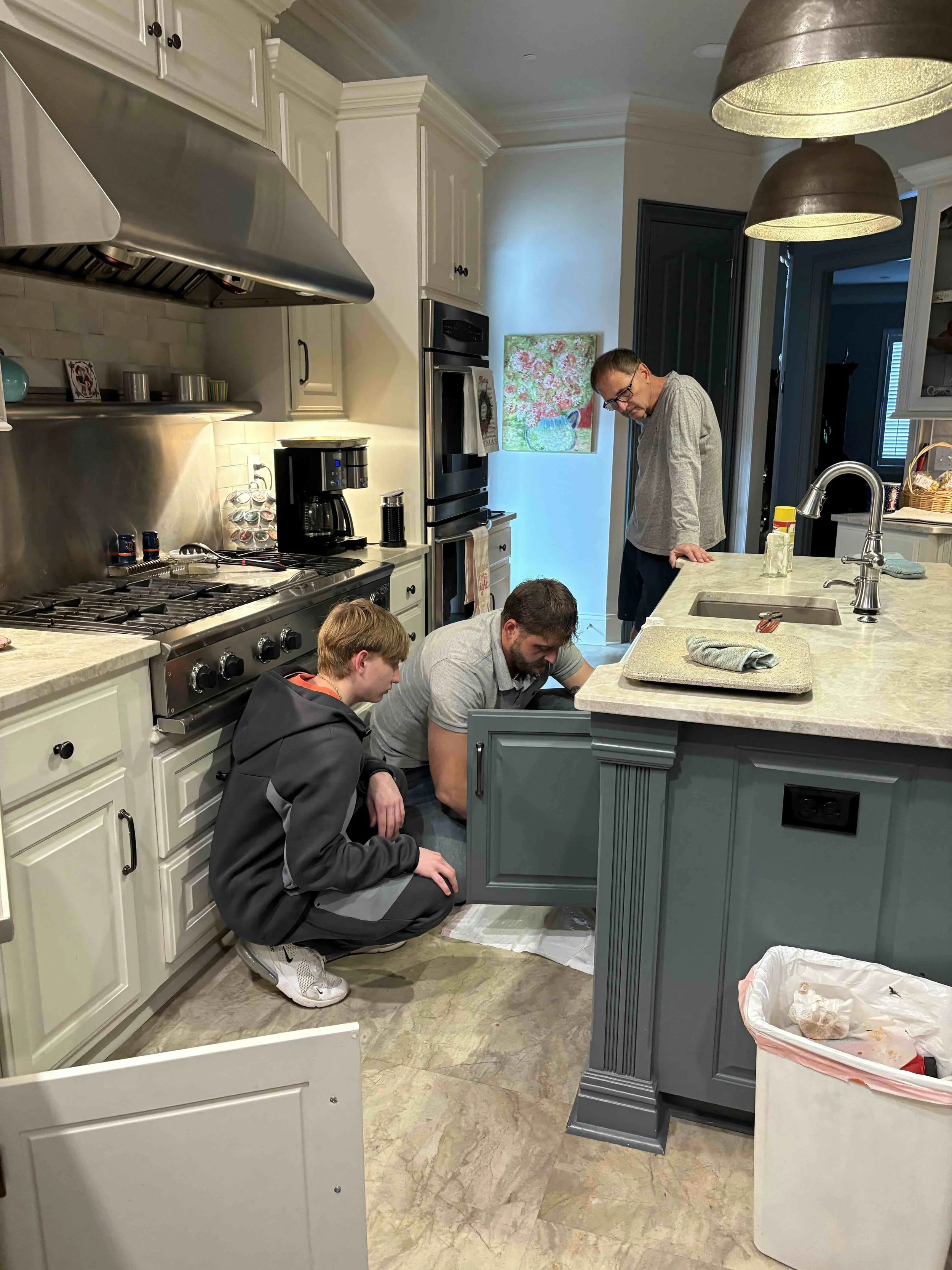 Three men in a kitchen, one kneeling under the sink inspecting it, another leaning over the counter, and a third standing nearby, all engaged in fixing or examining something under the kitchen sink.