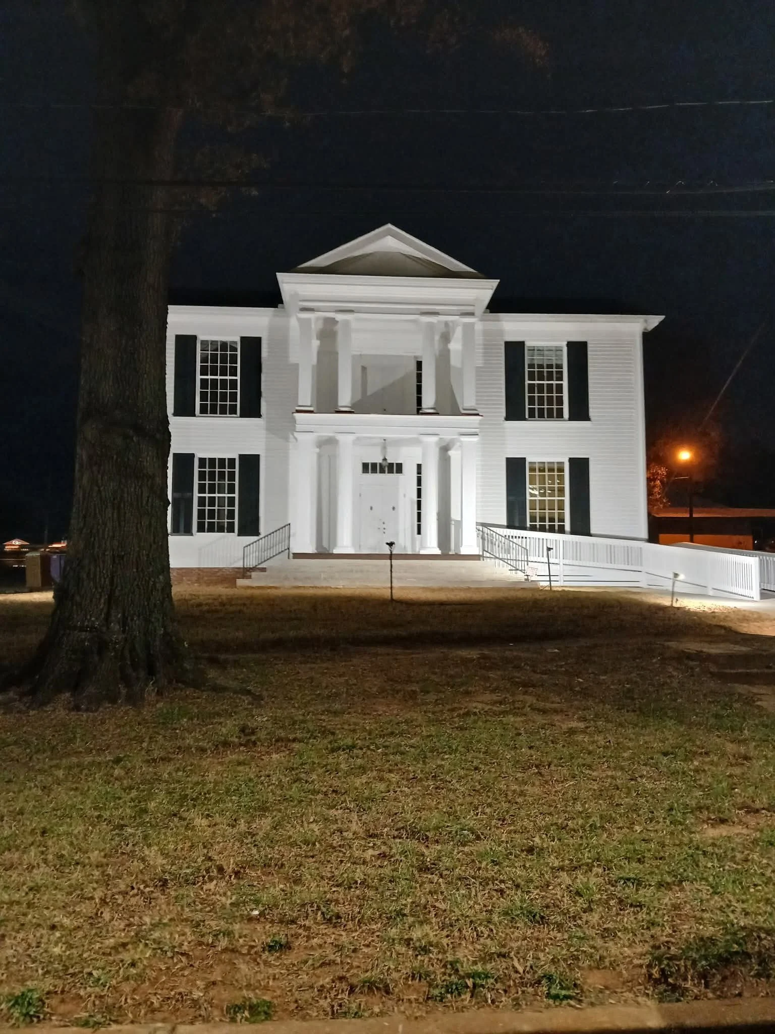 A white, large, two-story house with black shutters, tall columns, and stairs leading to the front door, illuminated at night.