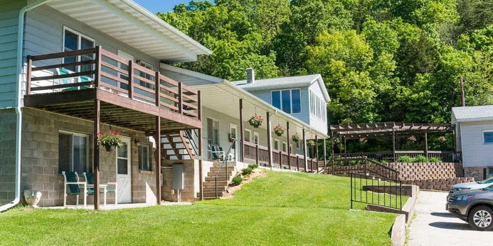 View of white motel rooms with brown decking, framed by green grass and mature trees beneath a clear blue sky.