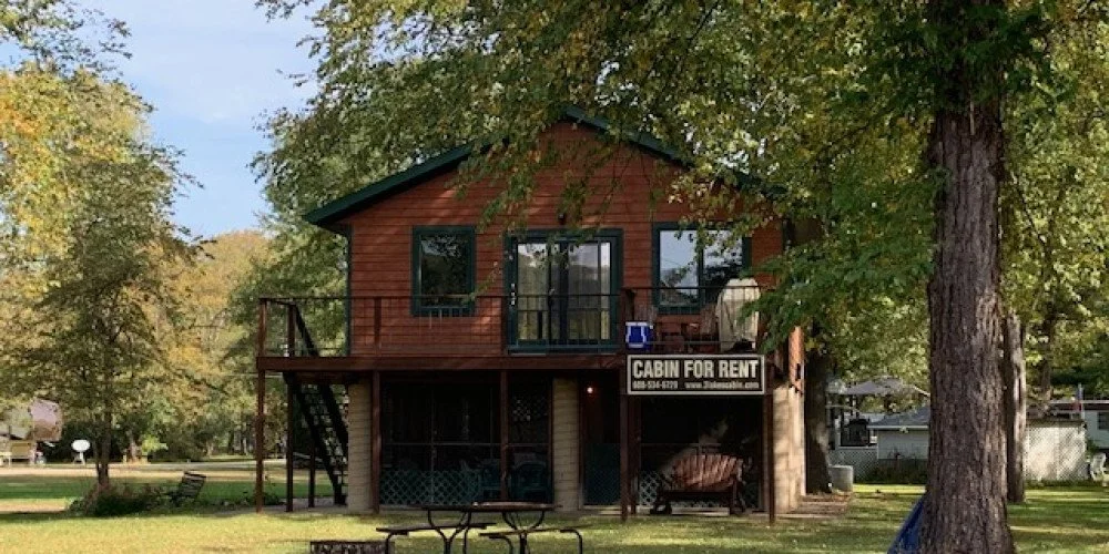 Brown cabin with green-trimmed windows and shutters, surrounded by lush green trees.