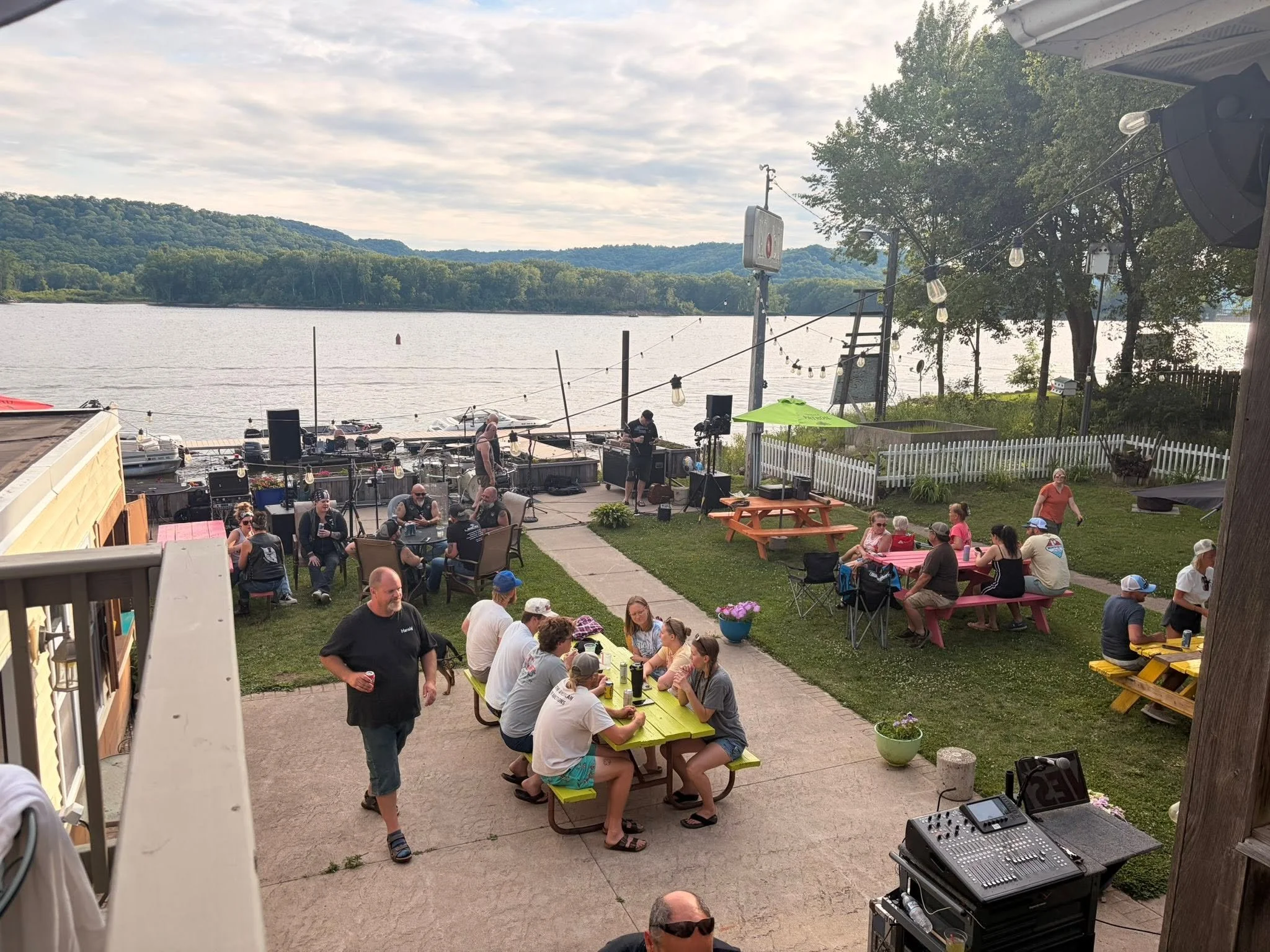 People gathering at Cat Daddy's Mississippi riverside patio with a view of water and bluffs in the background, some sitting at picnic tables and others standing, with musical equipment set up nearby.