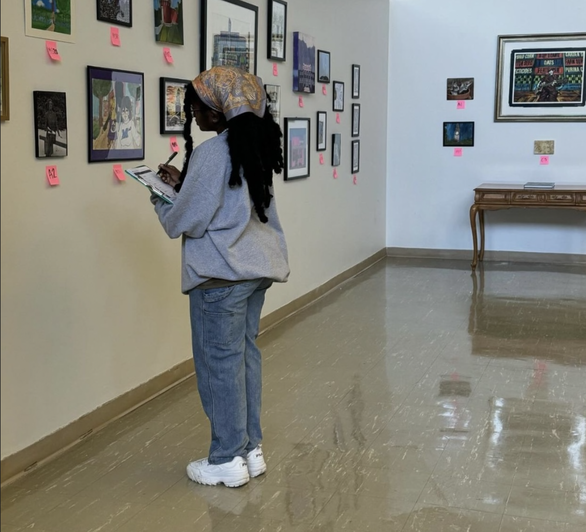 A woman with long braided hair, wearing a headscarf, glasses, a gray hoodie, blue jeans, and white sneakers, standing in an art gallery, holding a brochure and taking notes, looking at framed artworks on the wall.