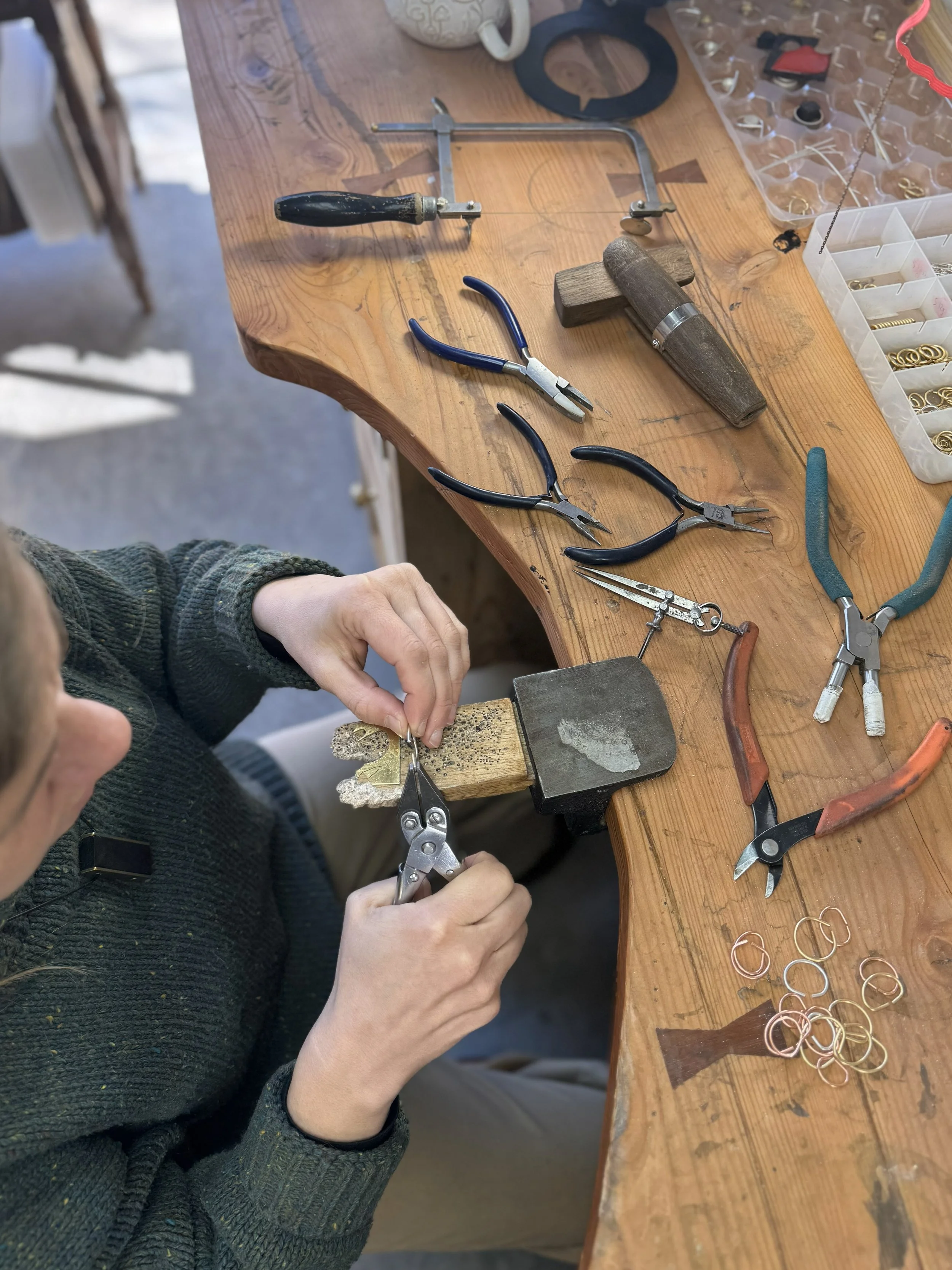A person working at a wooden workbench with jewelry-making tools, including pliers, cutters, a hammer, and rings, in a jewelry workshop.