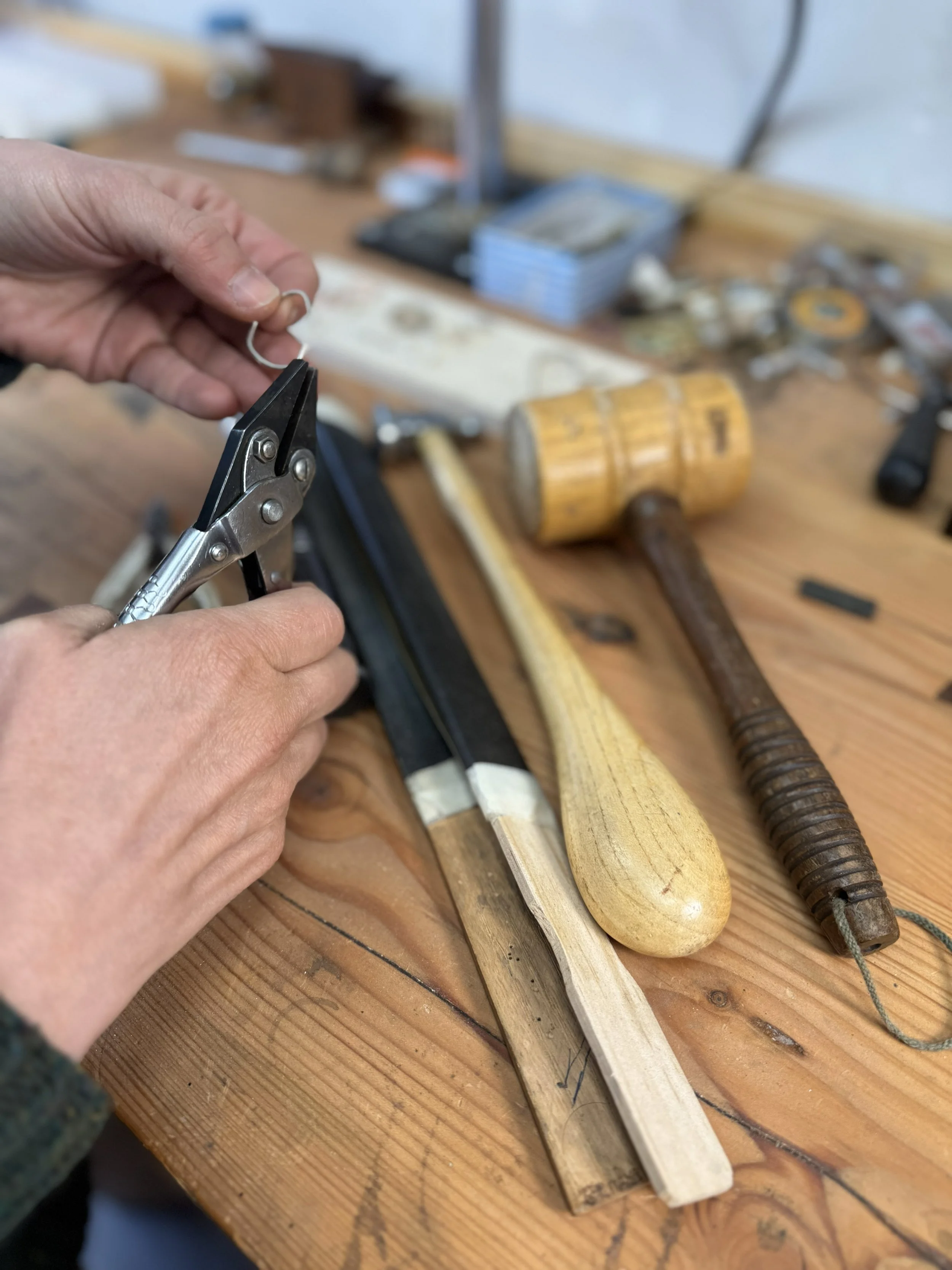Person using pliers on a small metal wire on a wooden workbench, with woodworking tools such as a hammer, a chisel, and various small hardware scattered on the surface.