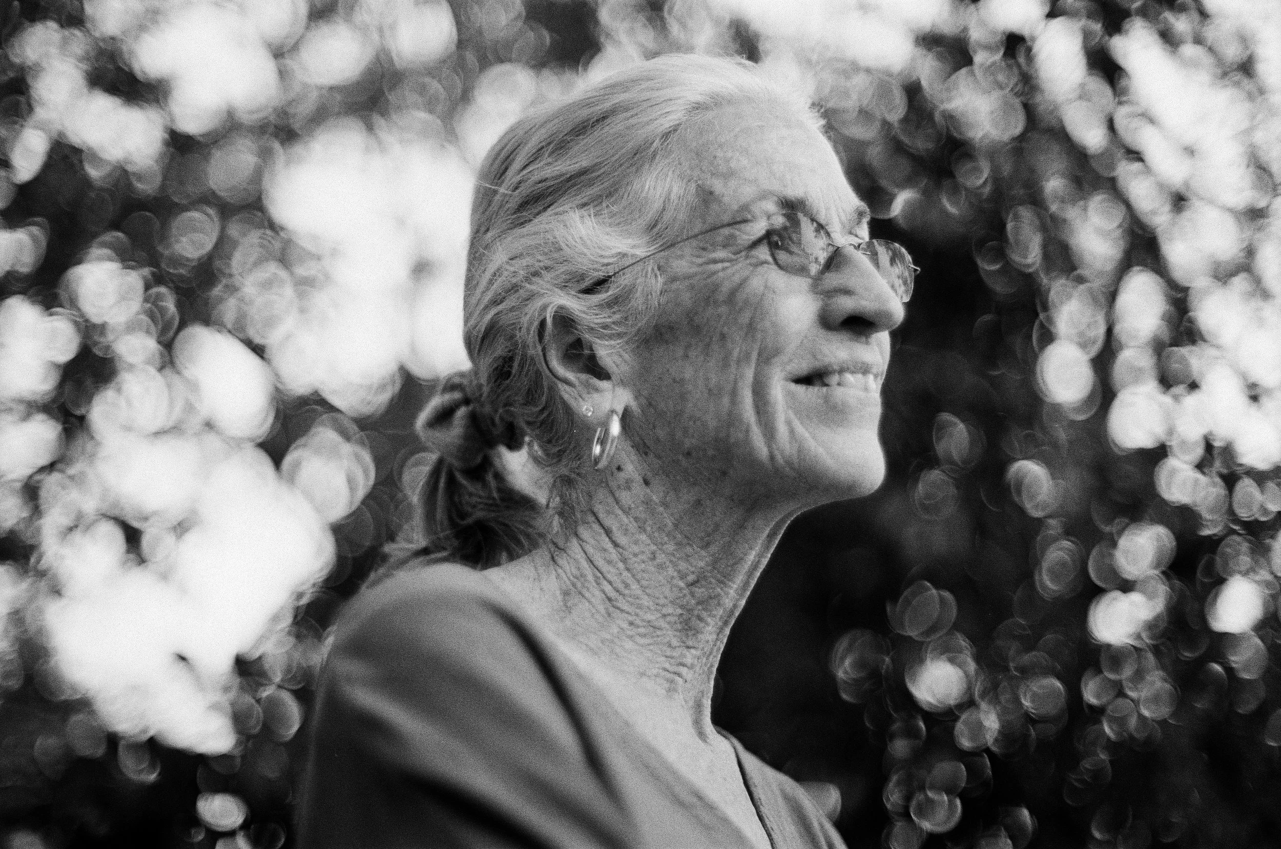 A black and white photo of an elderly woman with gray hair tied back, wearing glasses and earrings, smiling while looking off into the distance. The background consists of bokeh lights.