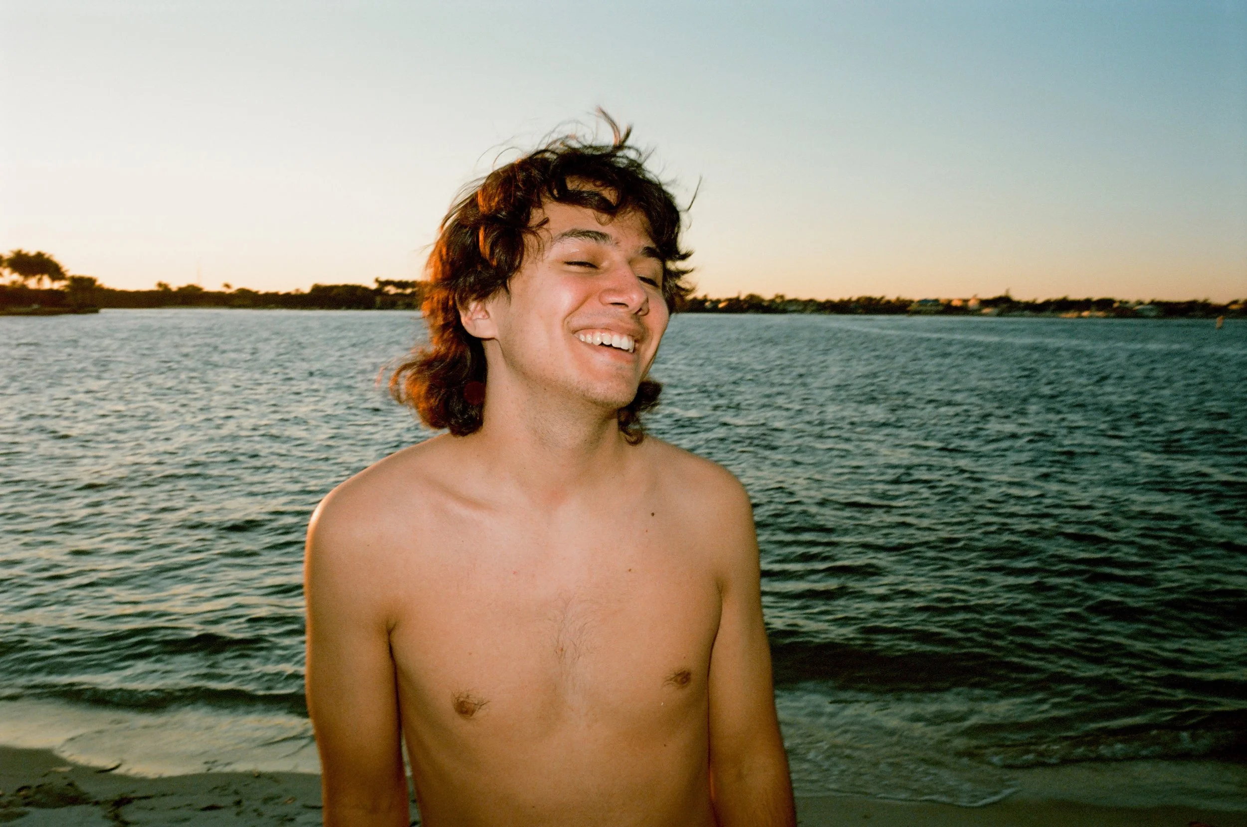 Young man smiling at the beach during sunset with water and shoreline in the background.