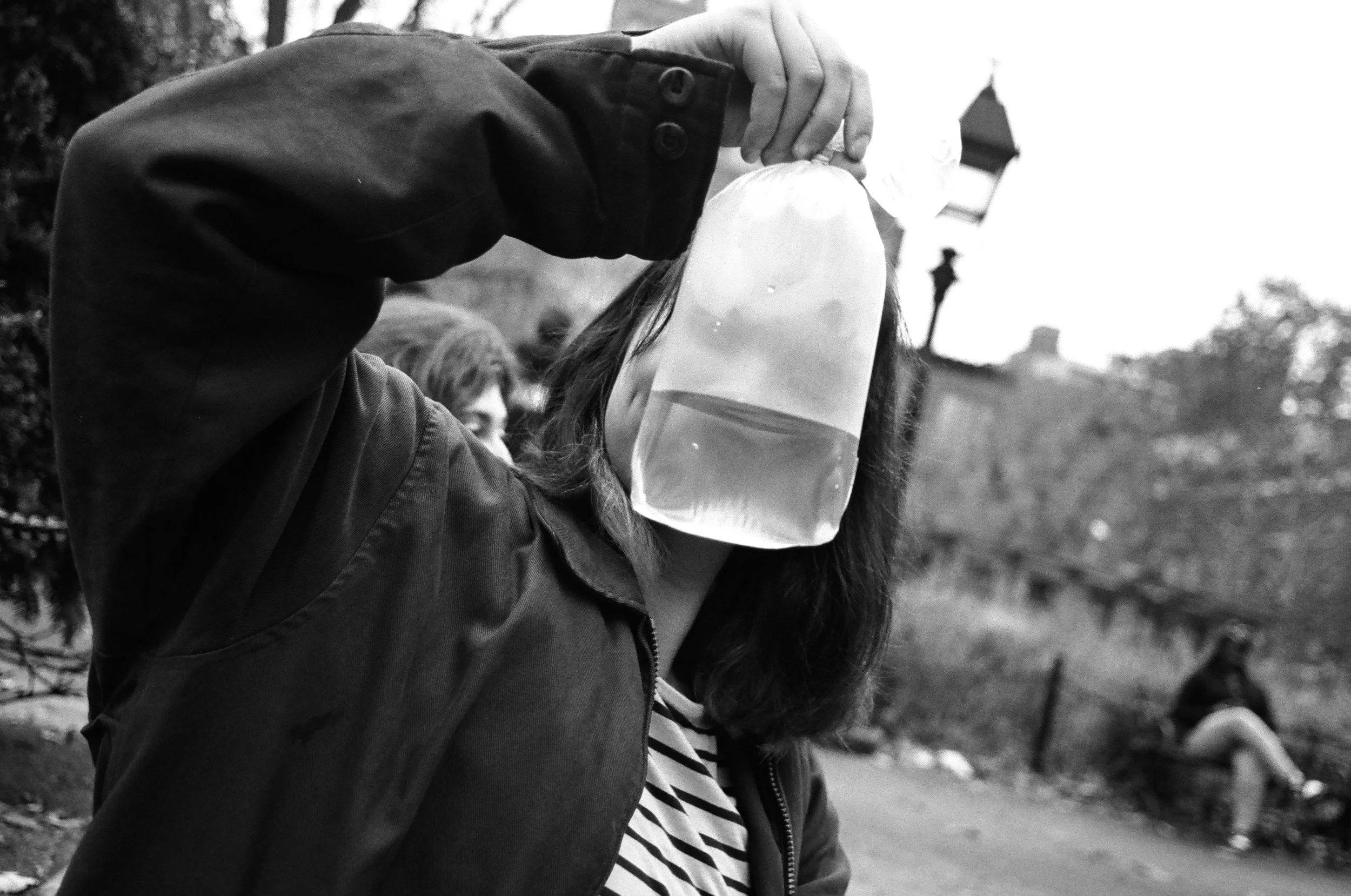 Person holding a large transparent water bag over their face outdoors in a park, with other people and trees in the background.