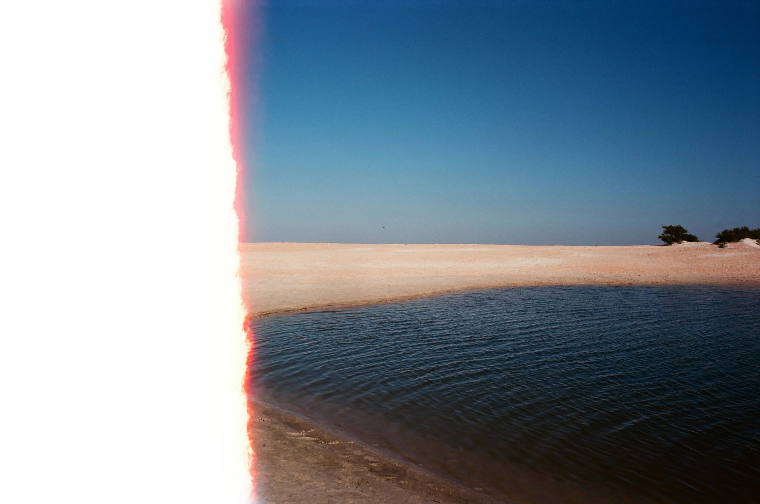 A landscape scene with a body of water in the foreground, a sandy shoreline, and a clear blue sky. The left side of the image appears damaged or corrupted with a white and red streak.