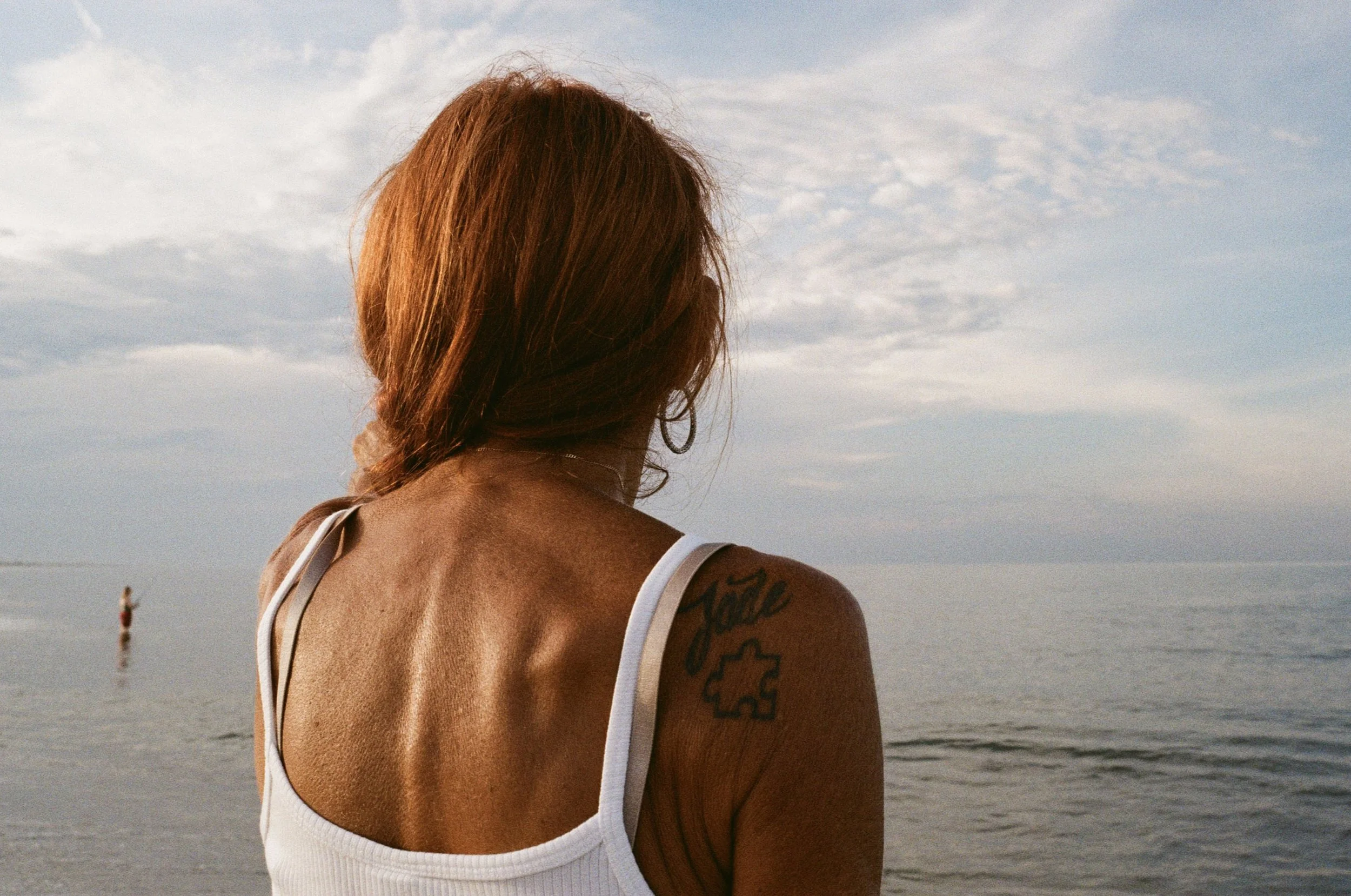 Woman with red hair looking at the ocean, wearing a white tank top, with a tattoo on her shoulder, partly overcast sky.