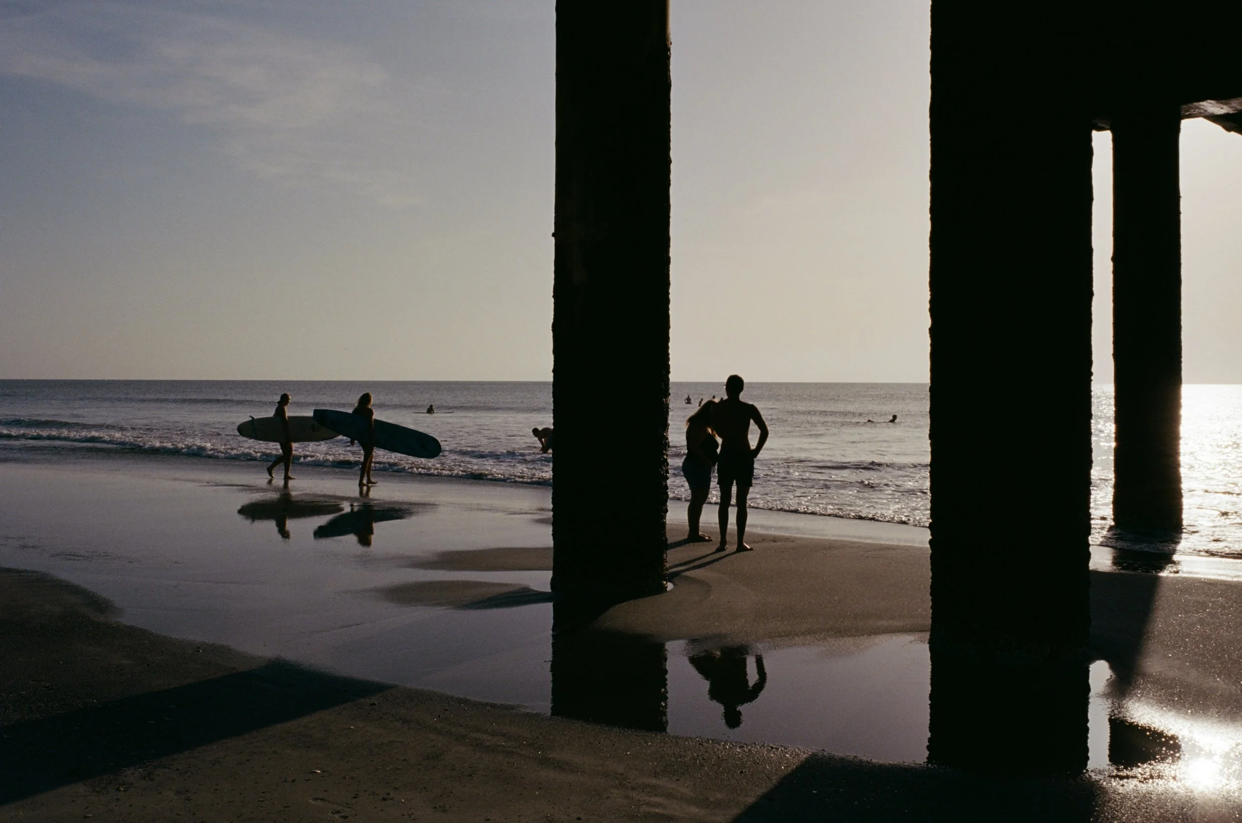 People at the beach near a pier during sunset, with some carrying surfboards and others walking along the shore, silhouetted against the light.