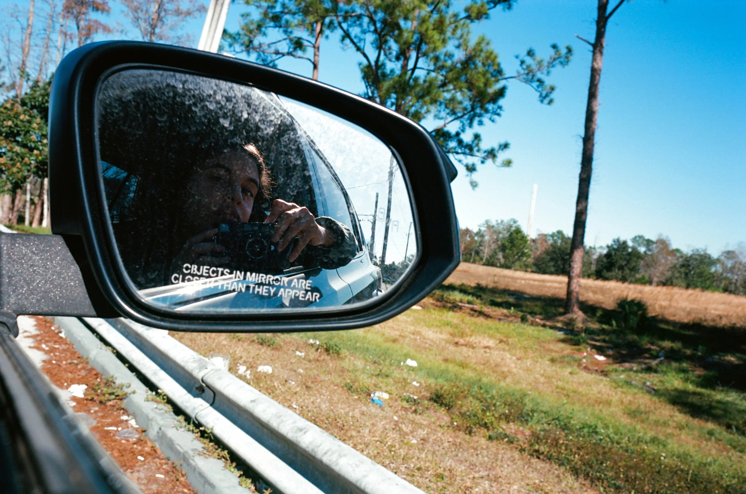 View of a woman taking a photo of herself in a car's side mirror, with a natural landscape and trees in the background.