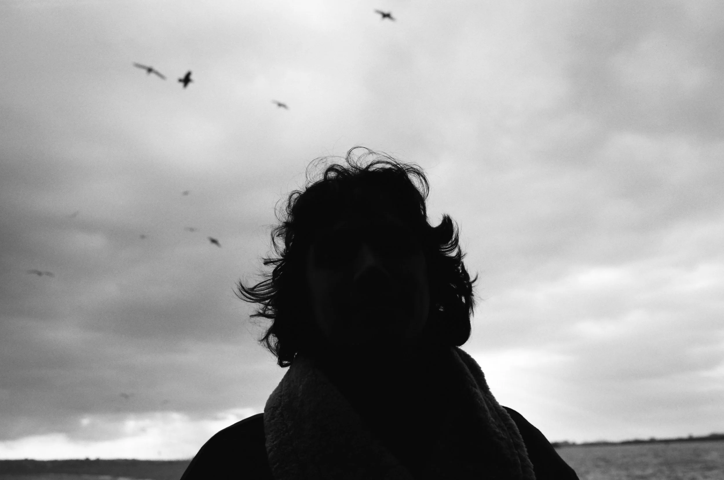 Black and white photo of a person with curly hair standing outdoors, with birds flying in the cloudy sky in the background.