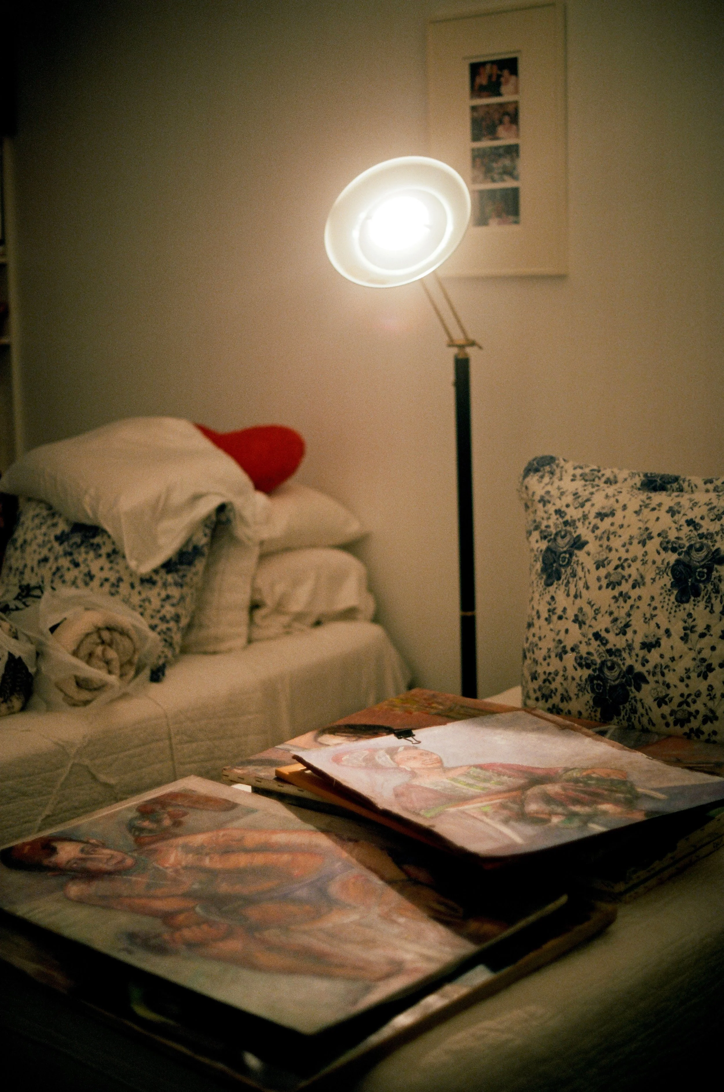 Indoor scene with a table, magazines, pillows, a sofa with blankets, a floor lamp, and framed pictures on the wall.