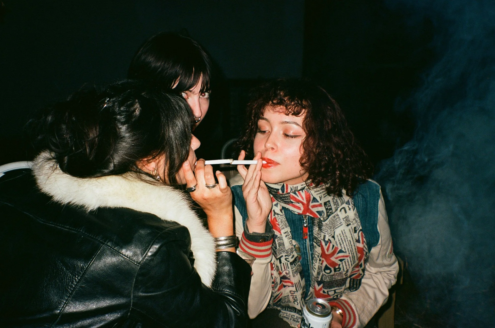 Three women at a party, one holding a cigarette and a soda can, another leaning in with a cigarette, engaging in a close conversation.